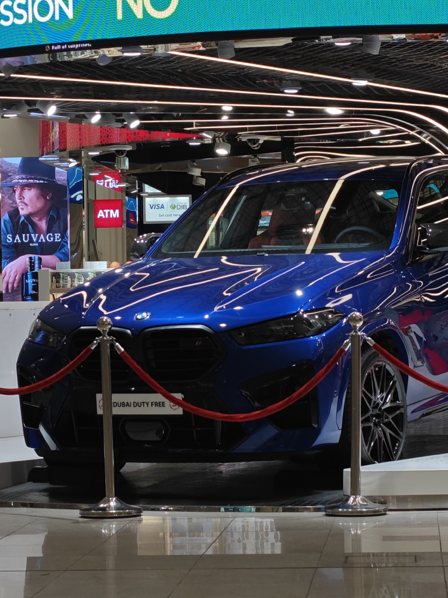 A blue BMW M5 is parked on display in a retail environment in Dubai, United Arab Emirates. The car is positioned behind a red rope barrier supported by silver stanchions. In the background, a large screen displays text and images, and a smaller display advertises Dior Sauvage perfume with a picture of Johnny Depp. An ATM machine is visible to the left of the car. The floor is tiled and reflective, and overhead lighting fixtures are present. The overall setting appears to be a duty-free shop in an airport or a similar retail establishment.