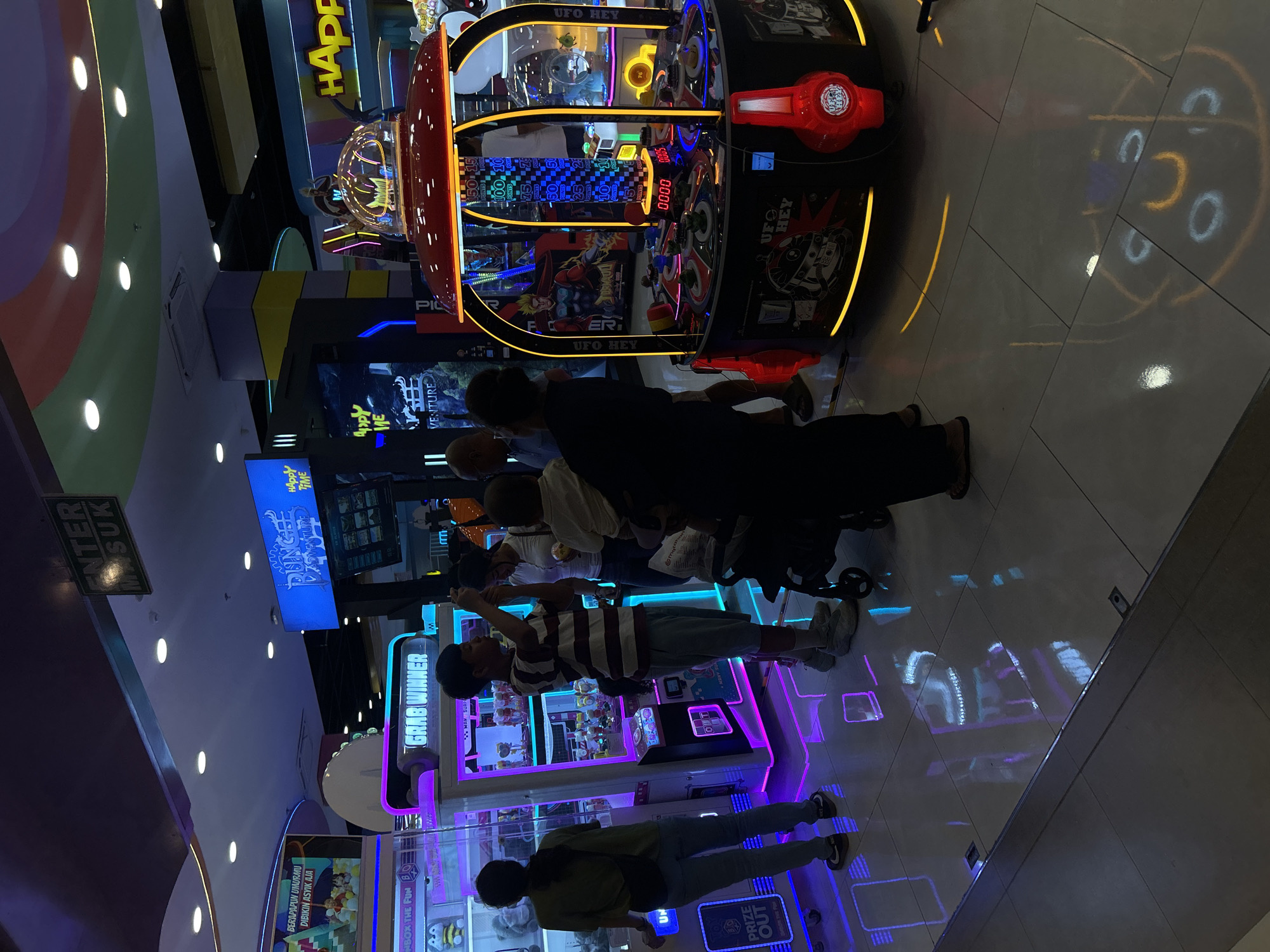 In Kuta, Indonesia, an indoor arcade, likely within a mall, is depicted with neon lights and reflections on the polished floor. Several people are visible, including a woman in a green shirt and jeans, a young boy in a striped shirt holding a camera, and an adult holding a baby. Other individuals are blurred in the background. The arcade features various game machines, including a claw machine labeled "GRAB WINNER" and a UFO-themed game with a digital score display. Signs for "Dunge Adventure" and "Happy Time" are also present. The atmosphere appears lively, with colorful lighting and the presence of entertainment attractions.