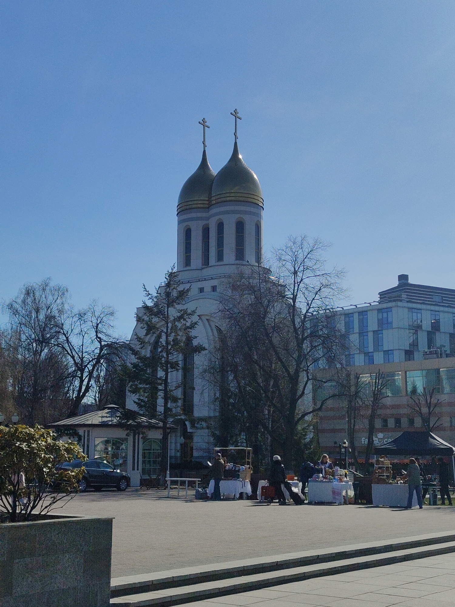 The image depicts a scene in a public space, likely a square, in the city of Unknown, Unknown. Dominating the background is a large Orthodox church with twin golden domes topped with crosses. The church is white with arched windows. In front of the church, there is an outdoor market or fair in progress. Several white tables are set up, displaying various items for sale. People are interacting with the vendors and browsing the goods. A black tent is also part of the market setup. Bare trees are scattered throughout the scene, suggesting either late autumn or early spring. To the left of the church, a smaller building or pavilion is visible, next to which a dark-colored car is parked. In the background to the right, a modern building with blue-tinted windows rises above the trees. The foreground consists of a paved area with steps leading towards the camera. A planter with a small bush is in the bottom left corner. The sky is a clear blue.