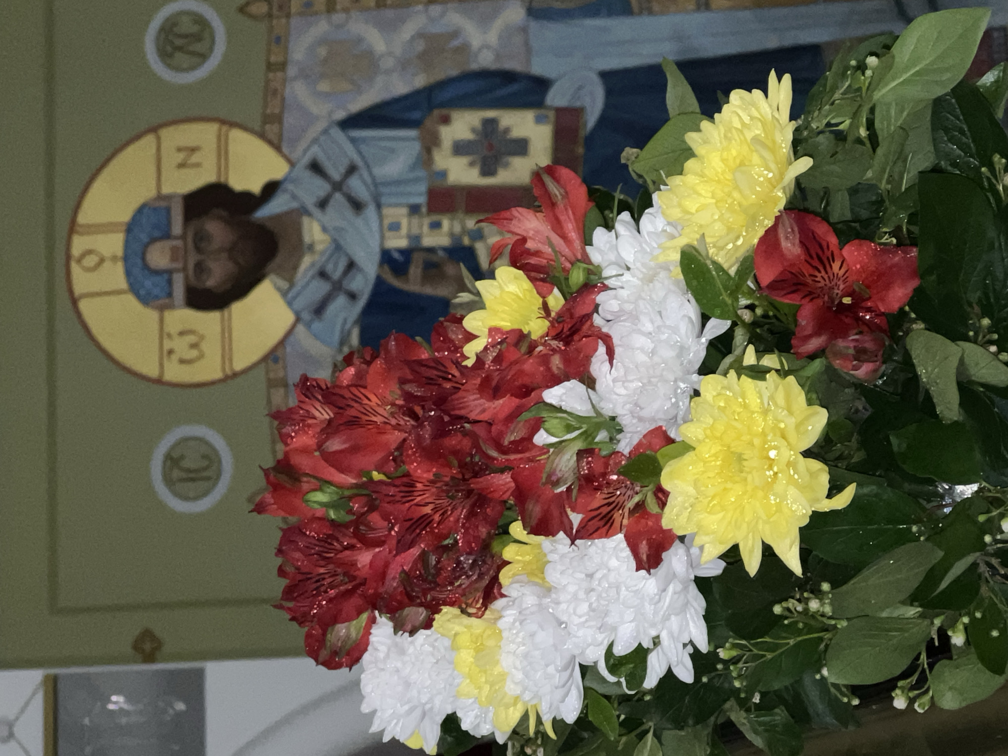 A bouquet of red alstroemeria, white chrysanthemums, and yellow dahlias is in front of a religious mural. The mural features a haloed figure with Greek letters around their head, and a blue and white cross-like design in the background. The location is possibly in a church in Khimki, Russia.