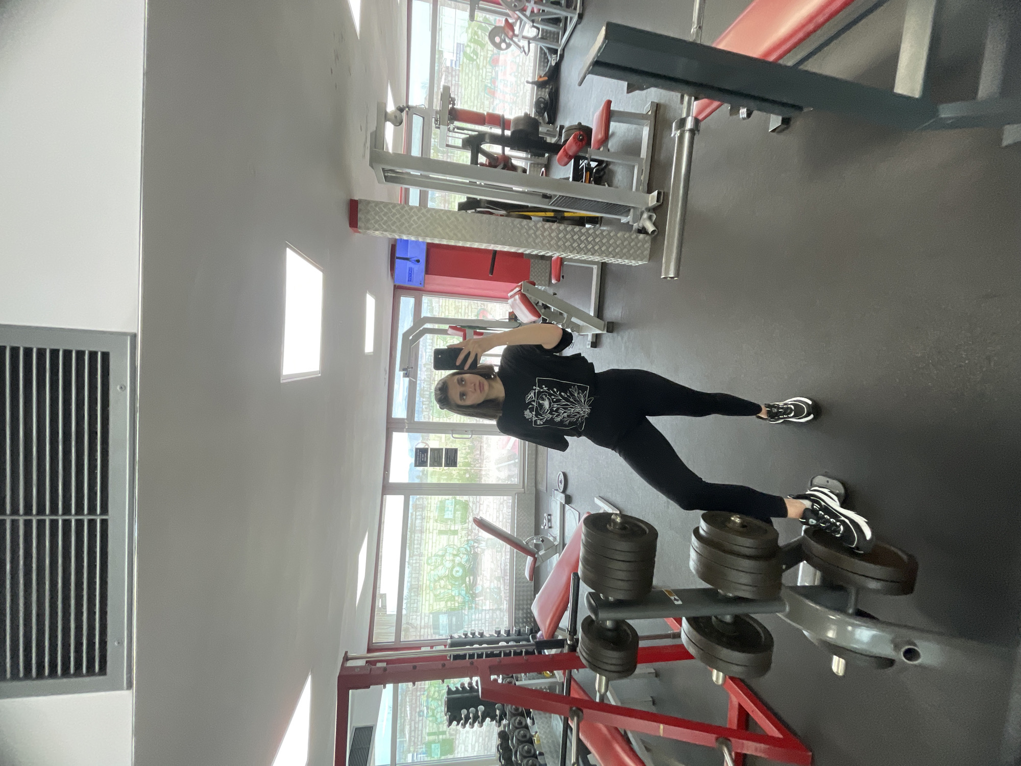 The image shows a young woman in a gym, taking a selfie. She is wearing black leggings and a black t-shirt. Her right foot is resting on the lower portion of the weight rack. The gym has several pieces of weightlifting equipment. There are also large windows. The location is likely in the city of Sofia, Bulgaria, as inferred from previous queries.