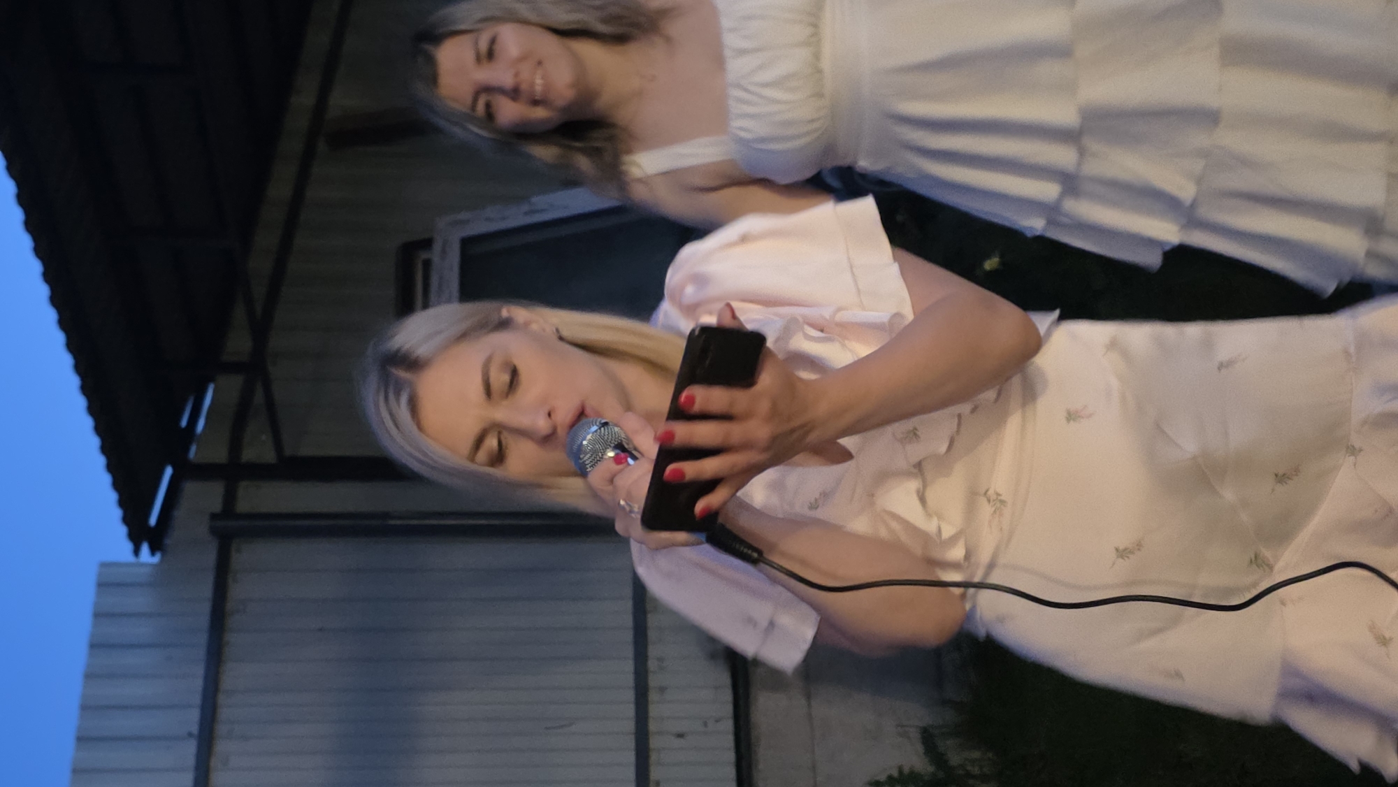 A woman with blonde hair and a pink dress with floral embroidery is holding a microphone to her lips and a smartphone in her hands. She is in the foreground, facing the viewer with her head tilted down. Behind her, a second woman with blonde hair and a white tiered dress is smiling. The scene appears to be outdoors, possibly in a backyard, with a corrugated metal fence and a dark awning in the background. The lighting suggests it is dusk. The location is identified as Bulgakovo, Russia.