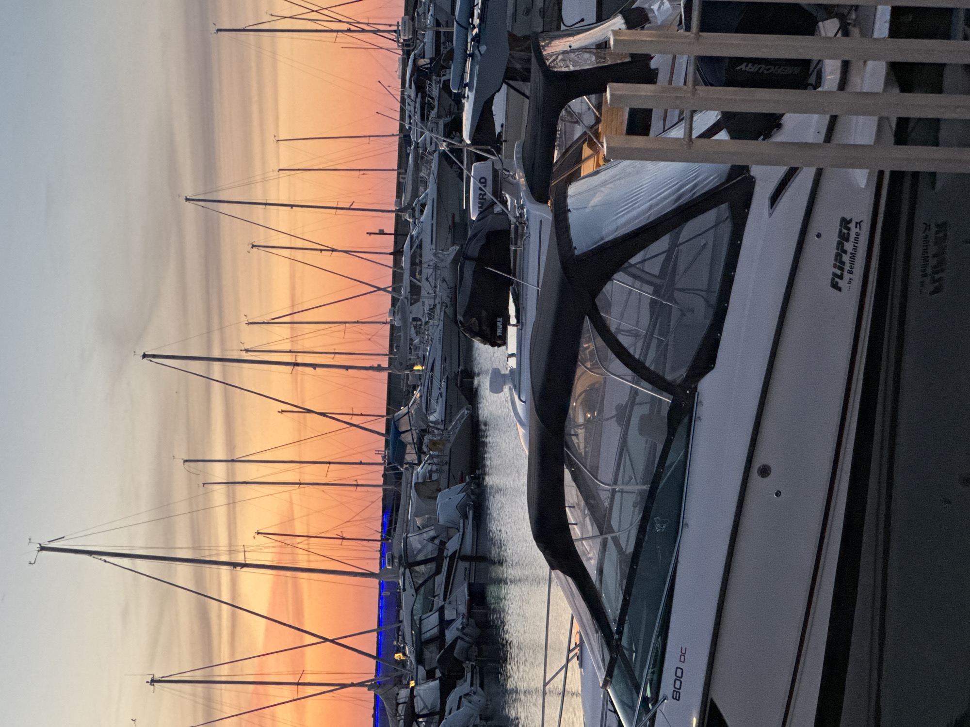 The image shows a marina at sunset, with numerous sailboats docked. The sky is a gradient of orange and pink, with hints of blue. In the foreground, a white motorboat with a black canopy is visible. The boat has "800 DC" and "FLIPPER ... by BellMarine" written on its side. The marina appears to be located in a city, with buildings and lights visible in the background. The water is calm, reflecting the sky and the boats. The overall atmosphere is peaceful and serene. The location is the Pirita Harbour in Tallinn, Estonia.