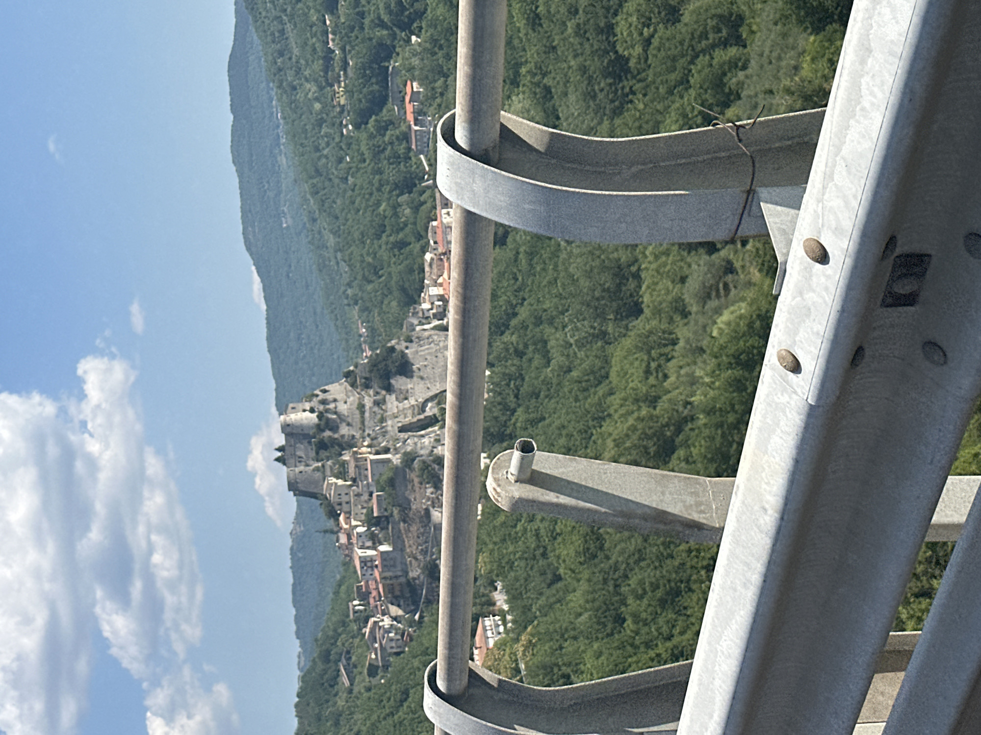 An eye-level, medium shot captures a view of the historic village of Cerro al Volturno, Italy, nestled atop a rocky incline and surrounded by lush, green mountains. In the foreground, a metal guardrail with gray posts partially obscures the scene. The village itself is a cluster of stone buildings with terracotta roofs, dominated by an ancient fortress with formidable stone walls and towers at its peak. The sky above is a vibrant blue, dotted with fluffy white clouds. The overall impression is one of picturesque historical architecture set against a dramatic natural landscape.