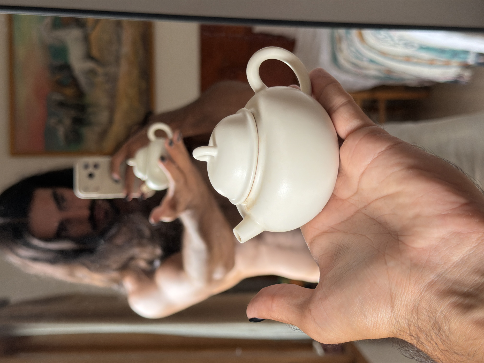 A person with long dark hair and a mustache is holding two small white teapots, one in each hand. The person is in the process of taking a photo of themselves in a mirror, holding up a smartphone in their left hand. The teapots are the focus of the image, with one in the foreground being held up in the person's right hand, and the other reflected in the mirror, held in the person's left hand. The background is blurred, showing a painting on the wall and a bed with a patterned blanket. The setting appears to be a bedroom. There is no indication that this image was taken in North Kuta, Indonesia.