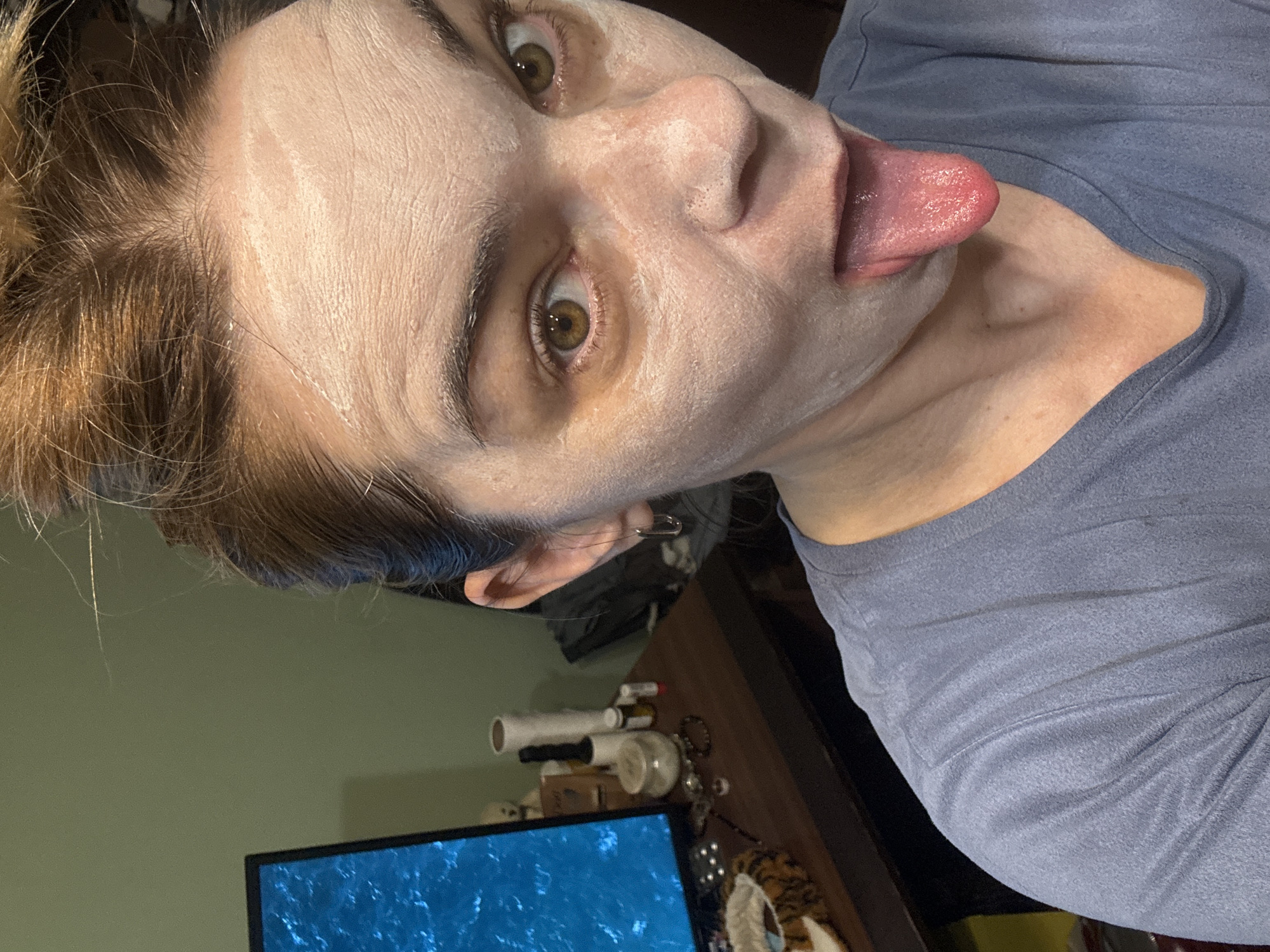 A person with light skin is facing the camera and sticking their tongue out. Their face appears to have a white cream or mask applied. They have light brown hair with darker roots. The person is wearing a gray-blue shirt. In the background, there is a computer monitor displaying blue water, and various objects are scattered on a dark-colored desk or table. The location is inside an unknown building in an unknown city.