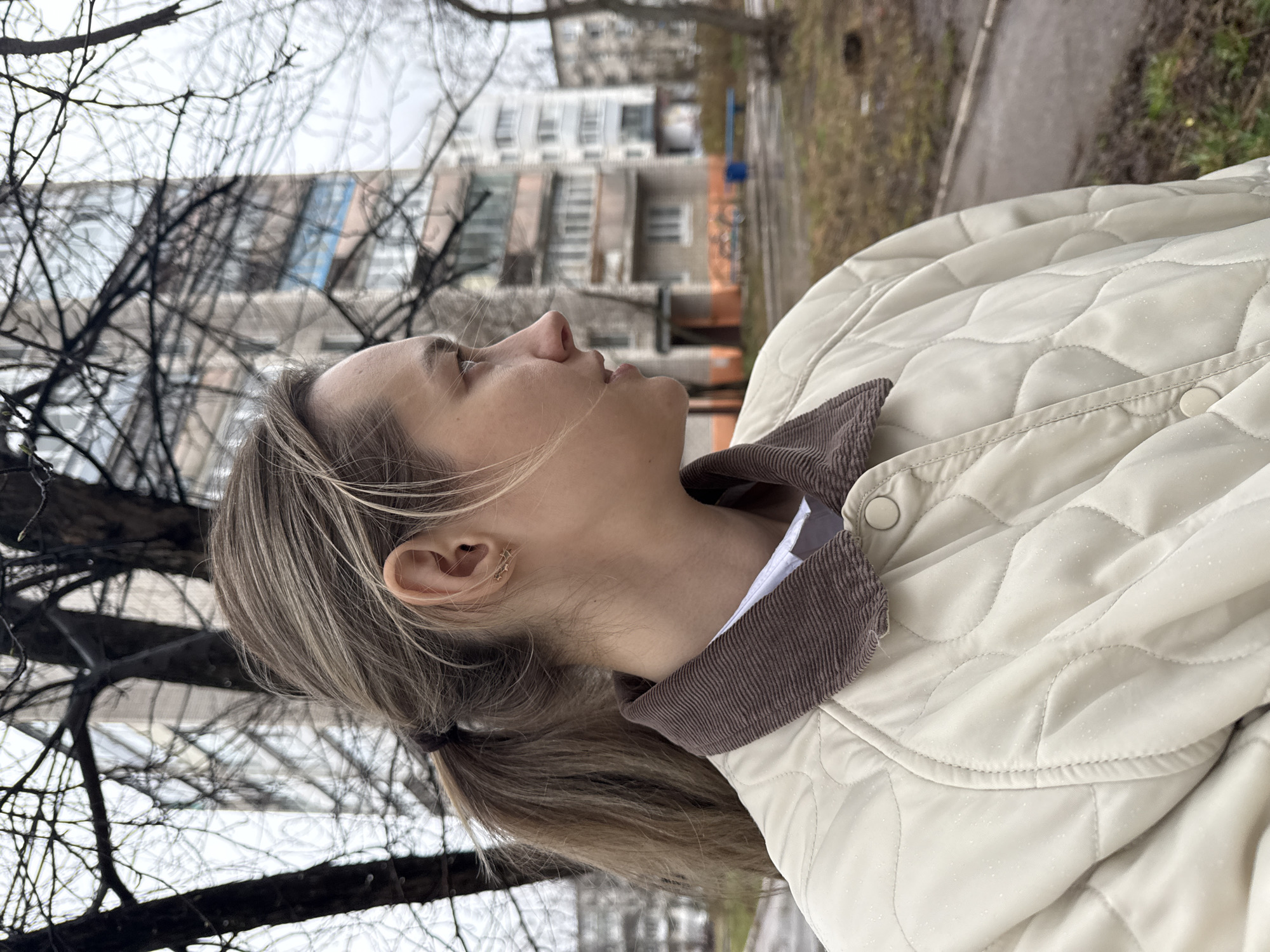 A profile view of a fair-skinned woman in her late 20s or early 30s, standing outdoors. She has light brown hair pulled back into a low ponytail. The woman is looking upward with a slightly open mouth. She wears a cream-colored quilted jacket with a brown corduroy collar. The background features bare trees and a multi-story building, indicative of an urban environment. Based on visual clues, this location may be in or similar to Chaikovsky, Russia. A sidewalk and some sparse vegetation are also visible.