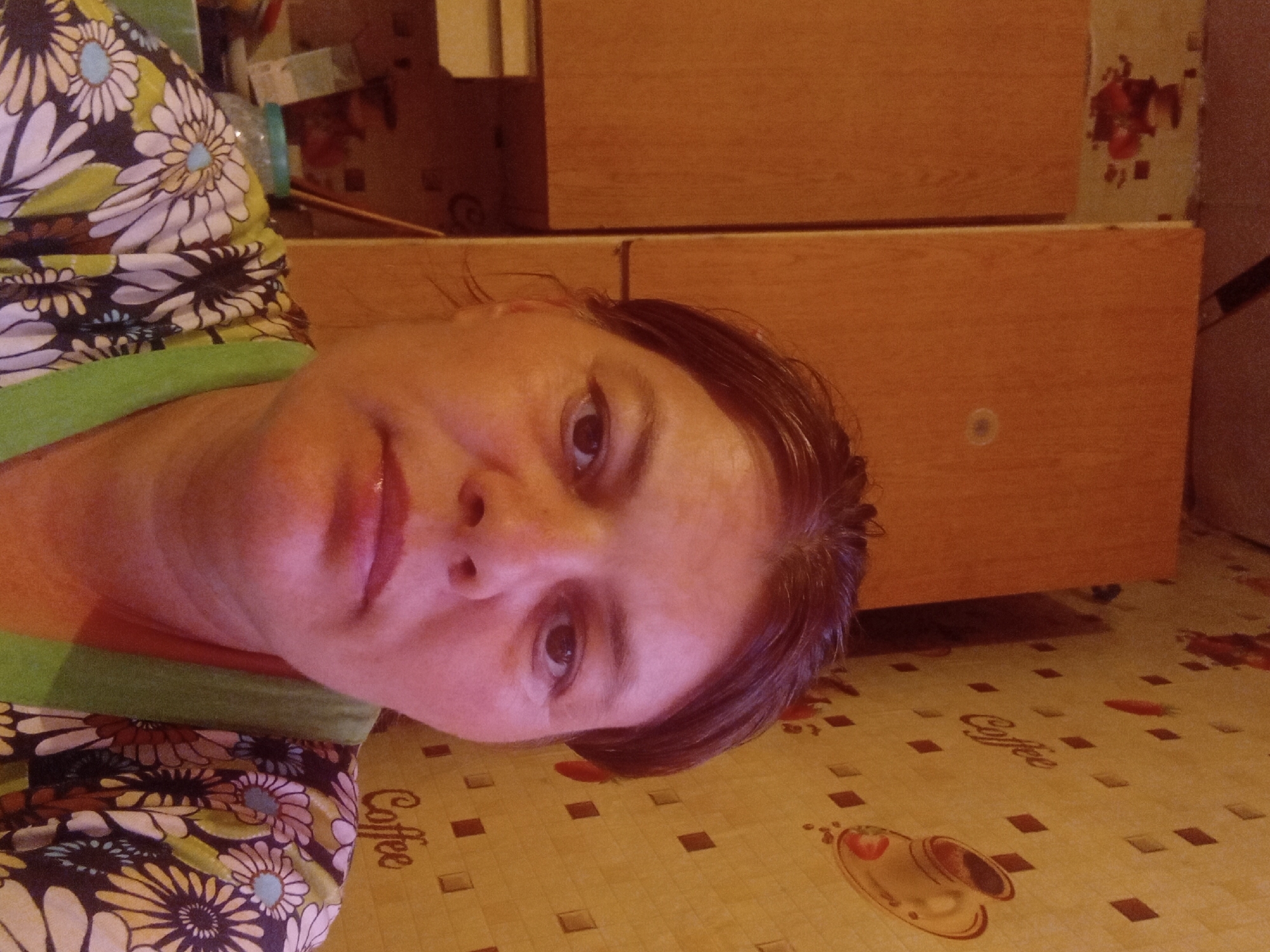 In a room with yellow patterned wallpaper depicting coffee cups and the word "Coffee", a woman with brown hair is taking a selfie. She is wearing a brightly colored floral shirt with a green neckline. Behind her, to the left, are two wooden cabinets. To the right, on the wallpaper, is a drawing of a coffee cup and saucer. The lighting in the room is warm and appears to be artificial. The location is the village of Malaya Vladimirovka, Malaya Vladimirovka rural district, Kazakhstan.