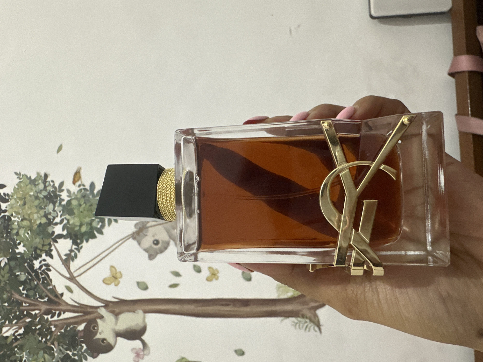 In Sukawati, Indonesia, a hand with pink-painted nails holds a clear glass perfume bottle. The bottle contains a dark amber liquid and is adorned with a gold YSL logo. The bottle is capped with a black rectangular lid, surrounded by a gold braided band. The background features a wall with a whimsical mural depicting a tree with cartoon cats and butterflies.