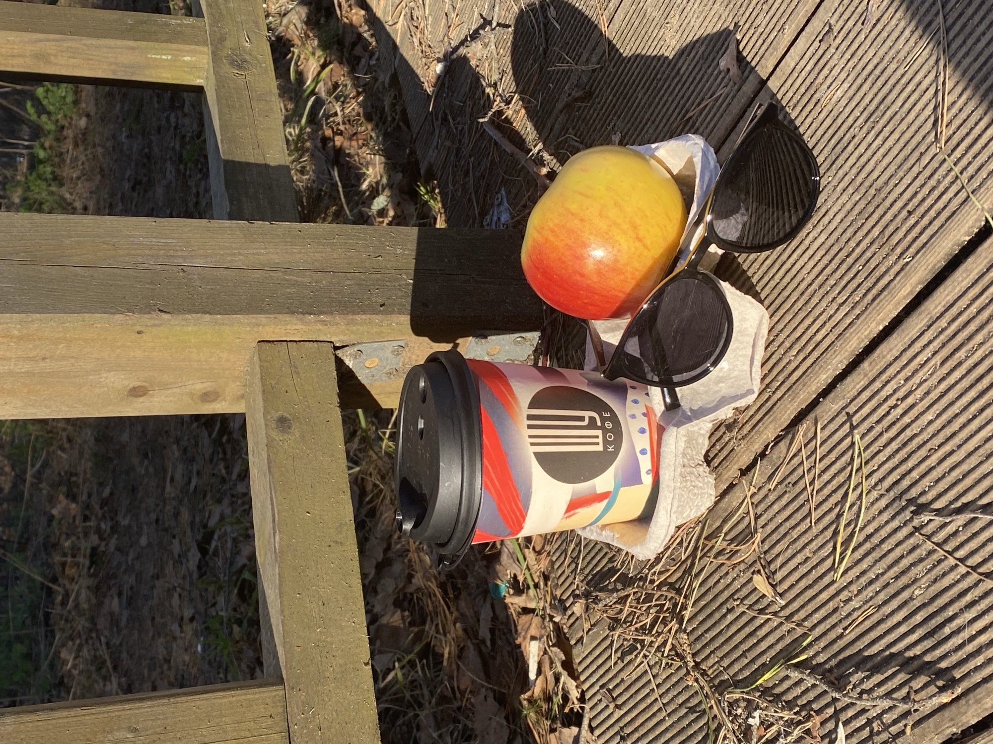 On a wooden deck or platform in what appears to be an outdoor space, a coffee cup with a black lid and a colorful patterned sleeve rests in a cardboard carrier. An apple, partially red and yellow, sits next to the cup, along with a pair of black sunglasses. Debris, including pine needles and small twigs, is scattered across the wooden planks. The deck is partially shaded, with sunlight illuminating sections of the wooden surface. A wooden structure, possibly a fence or steps, is visible in the periphery. The logo on the coffee cup contains the Cyrillic letters "КОФЕ" (coffee), indicating that the image was likely taken in a Russian-speaking environment, such as the city of Chaikovsky, Russia.