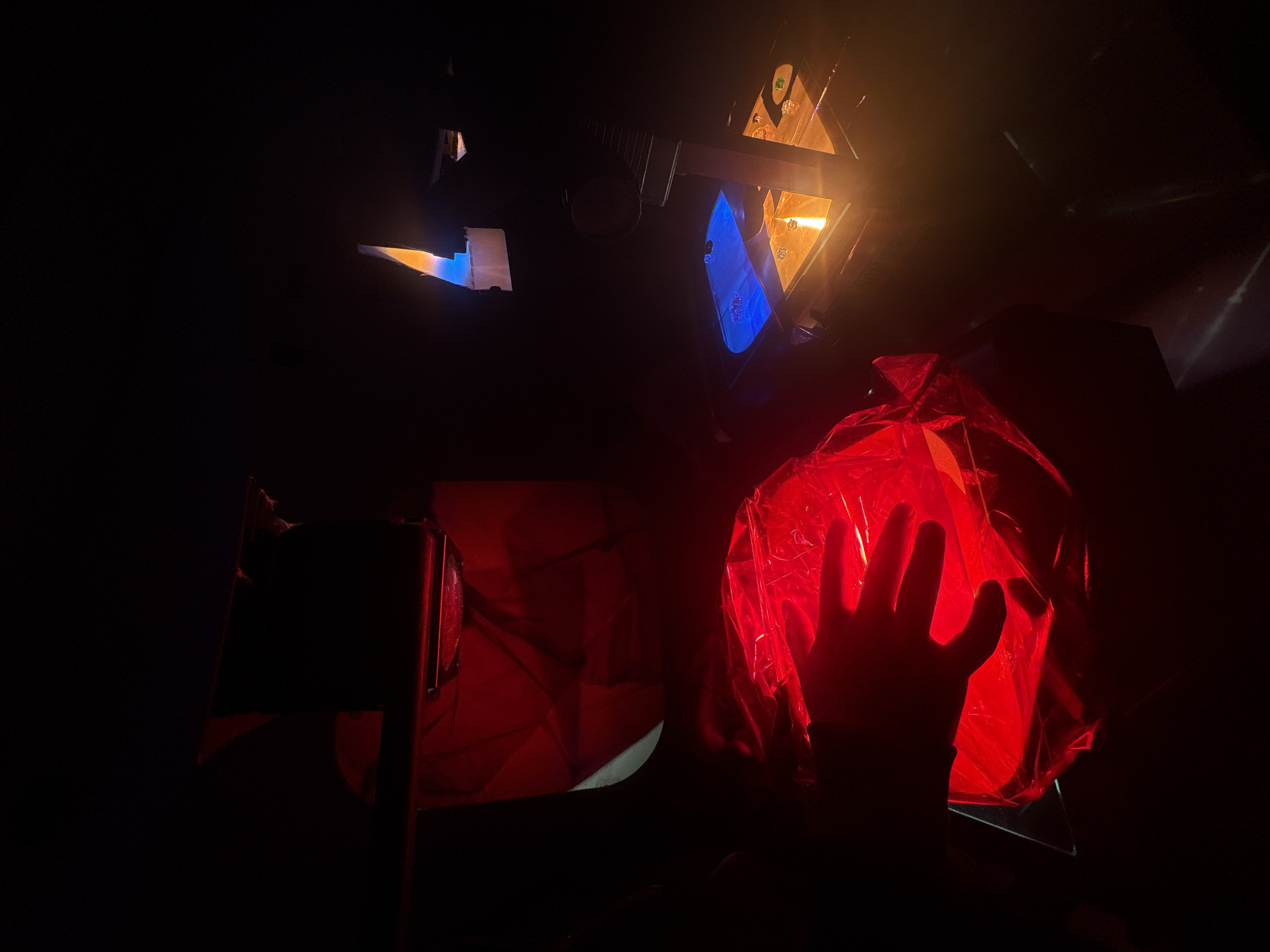 An indoor scene in Mexico City, Mexico, captured in low light conditions, is illuminated by multiple artificial light sources. In the lower-left, a human right hand, fingers spread, is silhouetted against and partially illuminated by a bright red light emanating from a crinkled, translucent red material. This material appears to cover a rounded object. Further back and to the left, a larger, angular structure is also bathed in red light. On the right side of the image, an apparatus features a small flame, exhibiting a blue base and an orange-yellow tip, positioned above a surface reflecting a vibrant blue light. Adjacent to the blue surface, another section of the apparatus reflects an orange-yellow light, possibly containing small, clear objects. A dark vertical support beam is visible as part of this apparatus. The background is predominantly dark, indicating a controlled lighting environment.