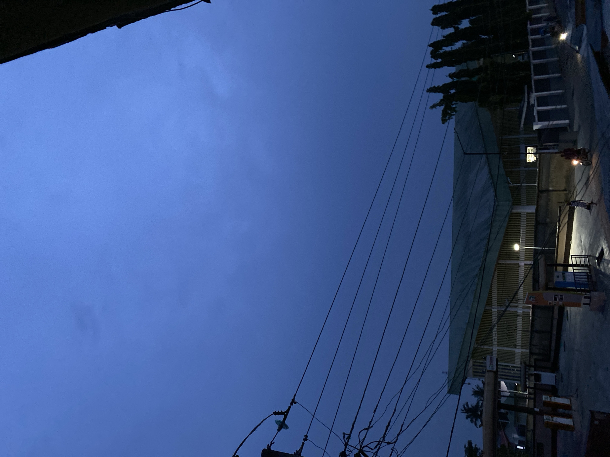 This image captures an urban or semi-urban street scene in Badagry, Nigeria, during twilight, likely dusk or dawn. The upper part of the frame is dominated by a deep, uniform blue sky with only faint traces of clouds, indicating a clear evening or morning.
A network of dark power lines and utility wires crisscrosses the mid-ground, extending diagonally from the lower-left to the upper-right of the image, supported by visible pole structures.
To the right, a long building or covered structure with a light-colored, possibly green-tinged roof is prominent. Artificial lights are active within the building and from a streetlamp positioned above it, casting a glow onto the surrounding area. Behind the building, a line of dark, silhouetted trees, including what appears to be a palm tree, adds to the local flora.
In the lower-right portion, a dark paved street or pathway runs alongside the building. A motorcycle with a brightly illuminated headlight is visible on this street, with a person faintly discernible nearby. There are also some unreadable signs on posts by the roadside. The combination of natural twilight and artificial illumination creates a serene yet active atmosphere, indicative of daily life transitioning between day and night.