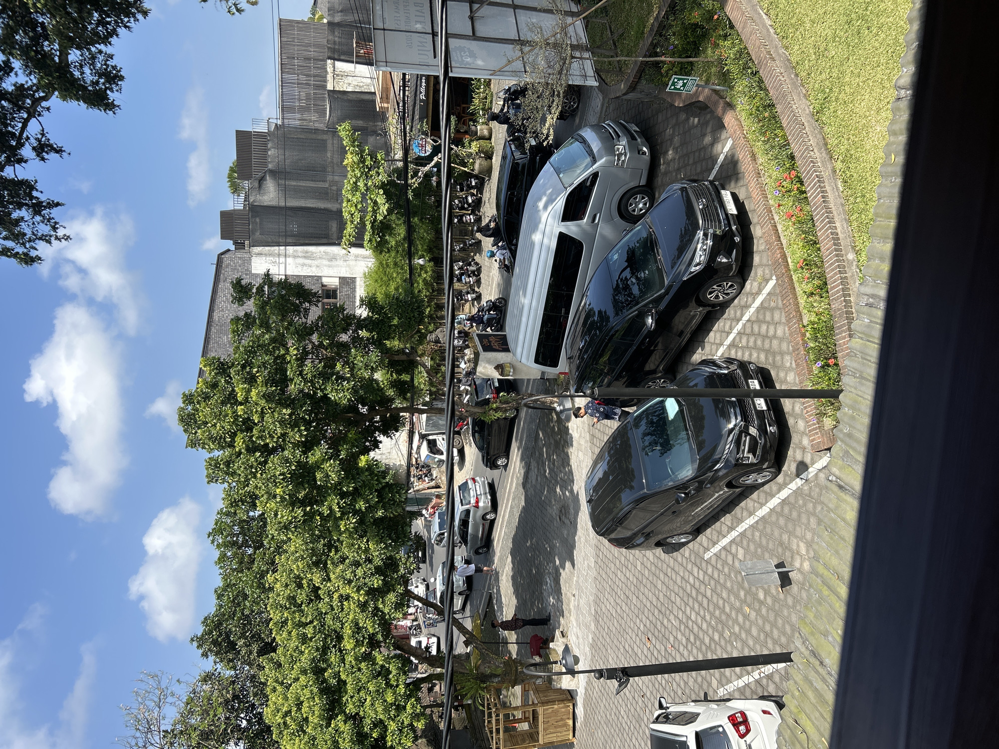 The image depicts a parking lot and a street scene in the Ubud District of Indonesia. In the foreground, a black SUV is parked on a paver surface. To its left, a white SUV is parked. Behind these vehicles, a silver minivan is parked, and to its right, another black SUV is parked. Several motorcycles are visible further back, with riders on them, suggesting movement. A street lined with trees and buildings stretches into the background. One building features a textured grey facade and modern architectural elements, with scaffolding or netting covering parts of it. The sky is blue with scattered white clouds. A person in a blue shirt is standing next to the silver minivan, and another person in a patterned shirt is standing near a bamboo structure in the parking lot. The overall scene captures a typical day in a bustling area, likely a commercial or tourist zone.