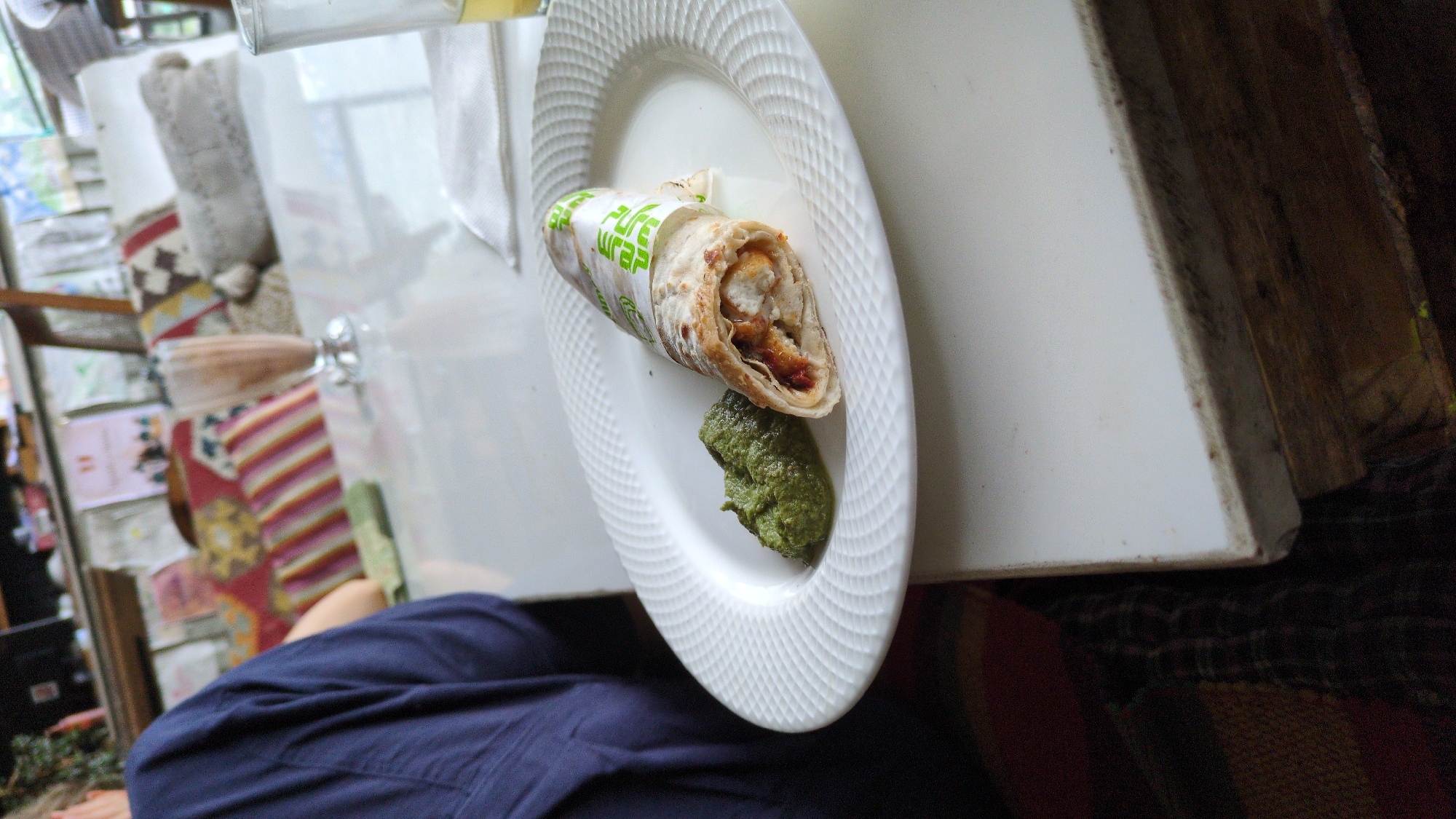 A tilted, medium-shot photograph shows a partly consumed wrap on a white plate with green sauce on a white surface. The wrap has a white and green wrapper with text. The white surface appears to be a table next to a dark wooden panel. In the background, there are cushions with various patterns and a shelf with books and other items. A person is partially visible, wearing a blue shirt. The location is Dharamkot, India.