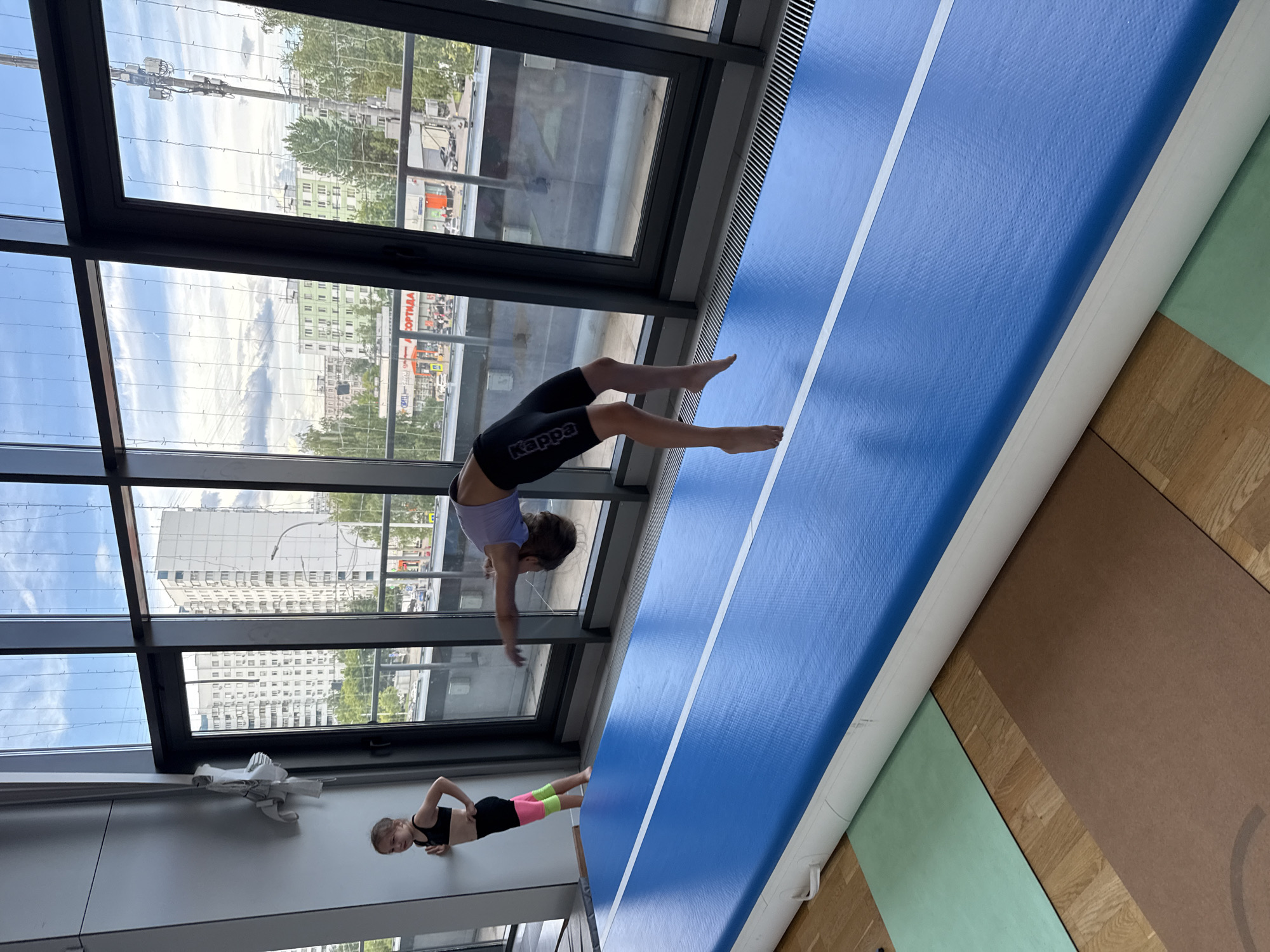 The image depicts two young girls in a gymnastics setting. The primary subject is a girl in mid-air, performing a gymnastic move that involves a handstand or cartwheel, with her legs extended. She is wearing a purple sports bra and black shorts with the "Kappa" logo visible. Her body is positioned horizontally, suggesting dynamic movement.
The second girl is visible on the lower left of the image, positioned on the floor. She is wearing a black sports bra and black shorts with colorful pink and neon green accents on the legs. She is in a push-up or plank-like pose, looking towards the camera with a neutral expression.
The setting appears to be an indoor gymnasium with large windows. Through the windows, an urban landscape is visible, featuring buildings and trees under a cloudy sky. The time of day seems to be daytime, given the natural light. The floor is a mix of wooden parquet and gym mats, including a prominent blue inflatable mat with white stripes, commonly used for gymnastics. There is also a light green mat and a brown mat visible. On the wall, there is a white object that resembles a folded piece of paper or a small sculpture.
The overall mood is one of active training and practice in a well-lit, spacious facility. The image is oriented in landscape, rotated 90 degrees counter-clockwise.