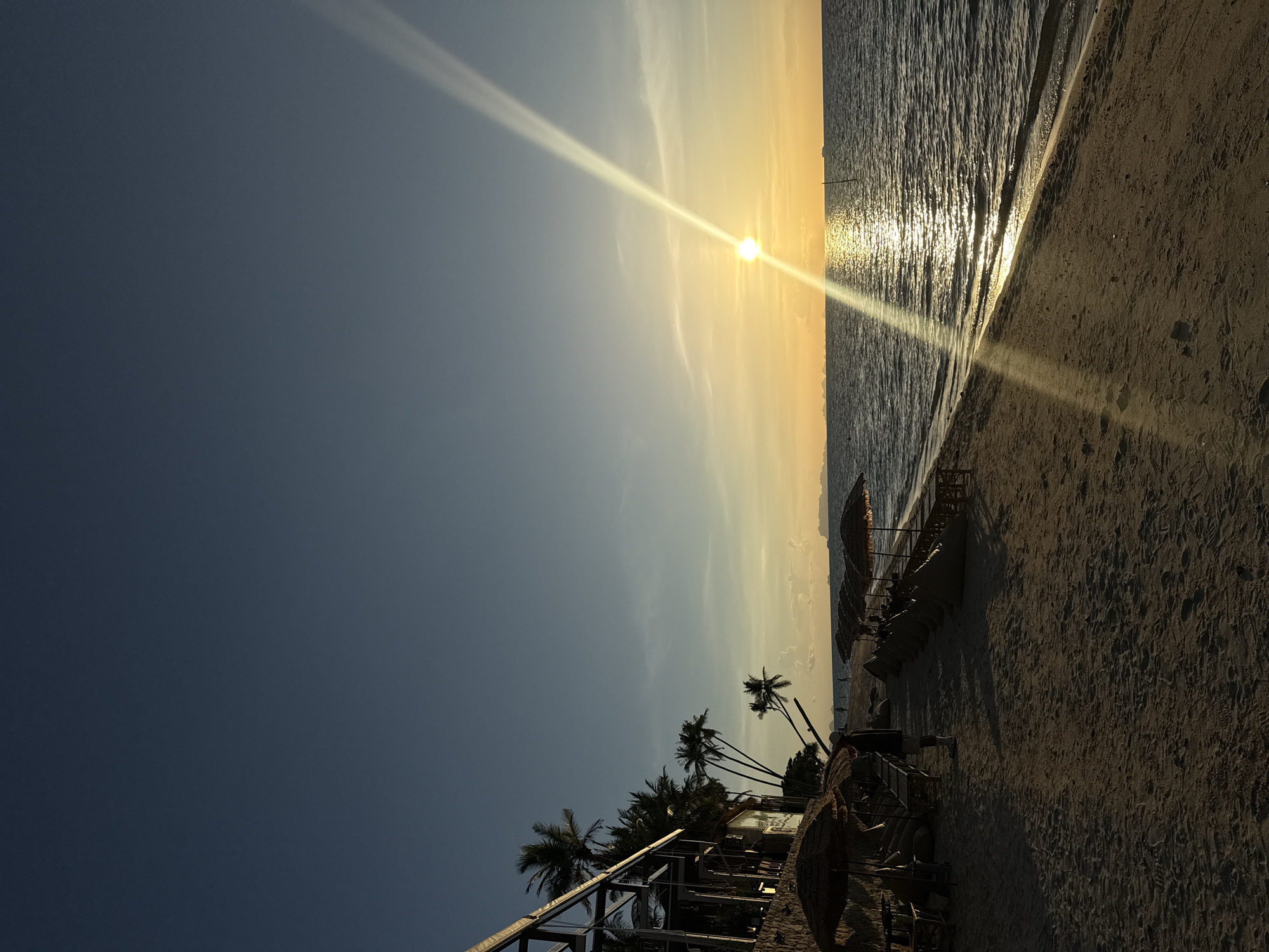 The image depicts a sunset scene at a beach in Baan Tai, Thailand. The sun is low on the horizon, casting a warm glow across the sky and reflecting on the water. The scene shows a sandy beach with rows of lounge chairs and umbrellas. Palm trees line the edge of the beach, and a building with a modern design stands on the left side of the frame. The ocean is calm, with gentle waves lapping at the shore. A single person is visible walking on the beach, and a few other figures can be seen in the distance. The sky is clear blue, with a few wispy clouds. The overall atmosphere is serene and picturesque.