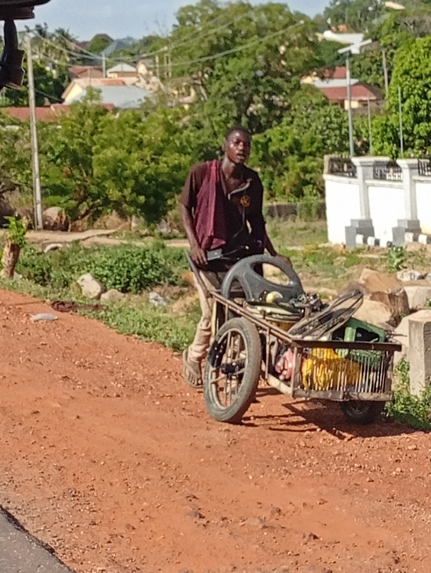 A man is pulling a heavily laden cart along a dirt road in an urban environment. The location is Unknown, Unknown. The man, who appears to be African, is wearing dark clothing and sandals. He is pulling a homemade metal cart that is loaded with various items, including what appears to be a bicycle wheel and a yellow object. The road is unpaved and has a reddish tint. In the background, there are trees, houses, and a white fence.