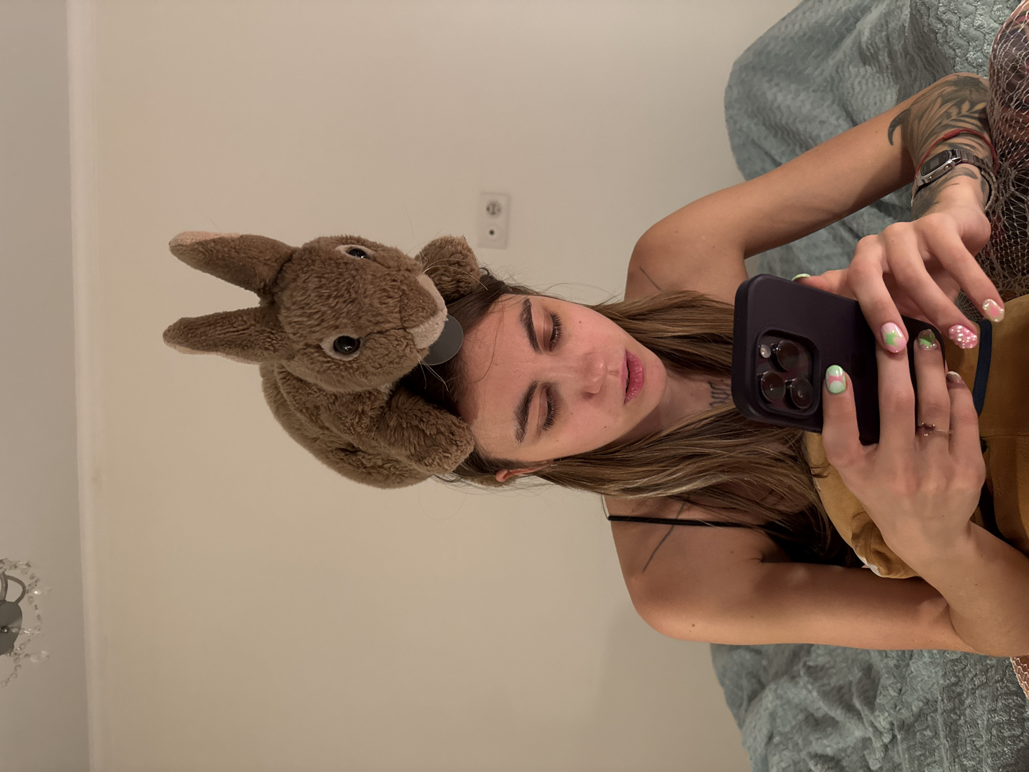 A young woman with long, light brown hair, wearing a black spaghetti strap top, is sitting on a bed with a teal textured blanket. A plush brown rabbit toy is perched on her head. She is looking down at her black smartphone, which she holds in her hands. Her nails are painted with a pink and green swirl design. She has tattoos on her arm and shoulder. The background is a plain, light-colored wall with a light fixture at the top and an electrical outlet with two sockets to the right of her head. The scene appears to be indoors, in a bedroom. The image does not contain any elements that allow for the determination of its location in Moscow, Russia.
