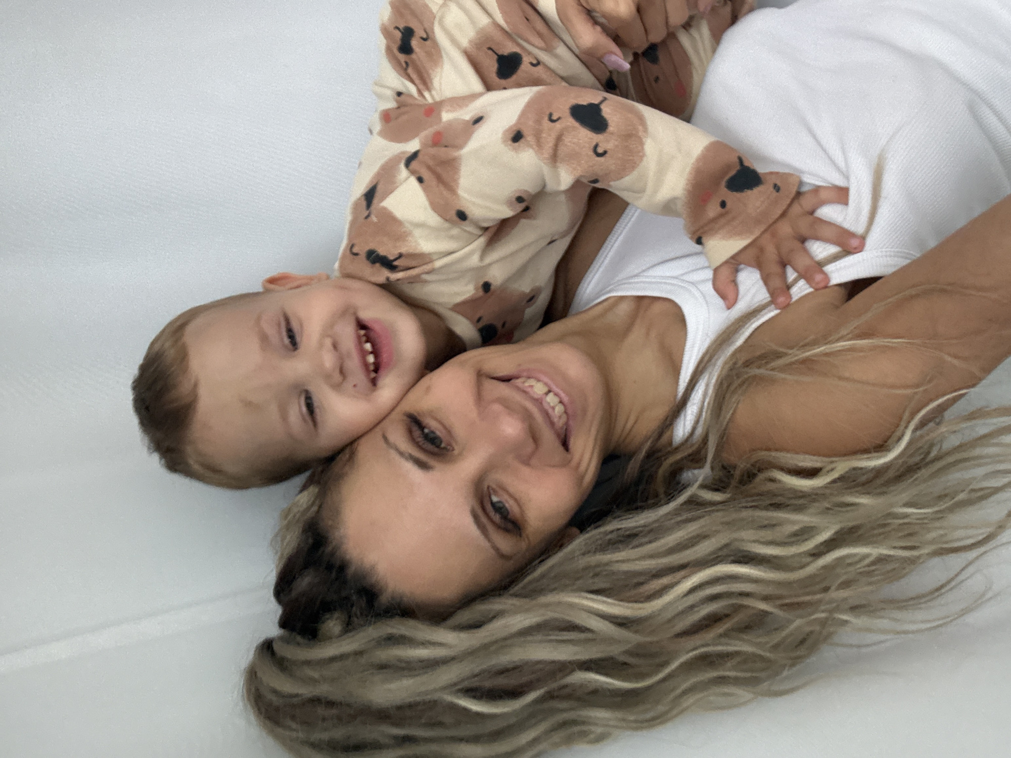 A woman with long, wavy blonde hair and a white tank top smiles as a toddler in a tan pajama set with a bear print embraces her. The child is resting on the woman's chest, with their arms around her neck. The background is a plain, light-colored wall. There are no discernible elements in the image that place the scene in Rusanovo, Russia.