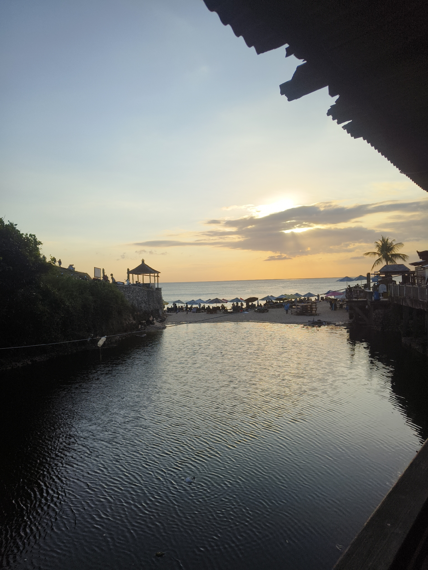 The image depicts a scenic view of a calm body of water leading to a beach with the ocean in the background, likely at sunset. The foreground is dominated by the dark, rippling water. To the left, a lush, green embankment rises, with a few silhouetted figures visible on its edge. Atop the embankment, a stone wall supports a small, traditional-looking gazebo.
Beyond the embankment, a sandy beach stretches out, dotted with numerous beach umbrellas and tables, suggesting a resort or dining area. More silhouetted people are scattered across the beach and near a wooden structure with a thatched roof on the right side of the frame, which appears to be part of a building or boardwalk. A palm tree stands near this structure.
The sky is a gradient of soft blues and yellows, with the sun partially obscured by clouds, casting rays of light. The overall ambiance is serene and tropical, characteristic of Kuta Selatan, Indonesia, particularly a location like the area near Uluwatu Temple, which features cliffside views and beaches. The water in the foreground could be a natural lagoon or a canal leading to the sea. The presence of umbrellas, tables, and a gazebo indicates a place for leisure and relaxation.