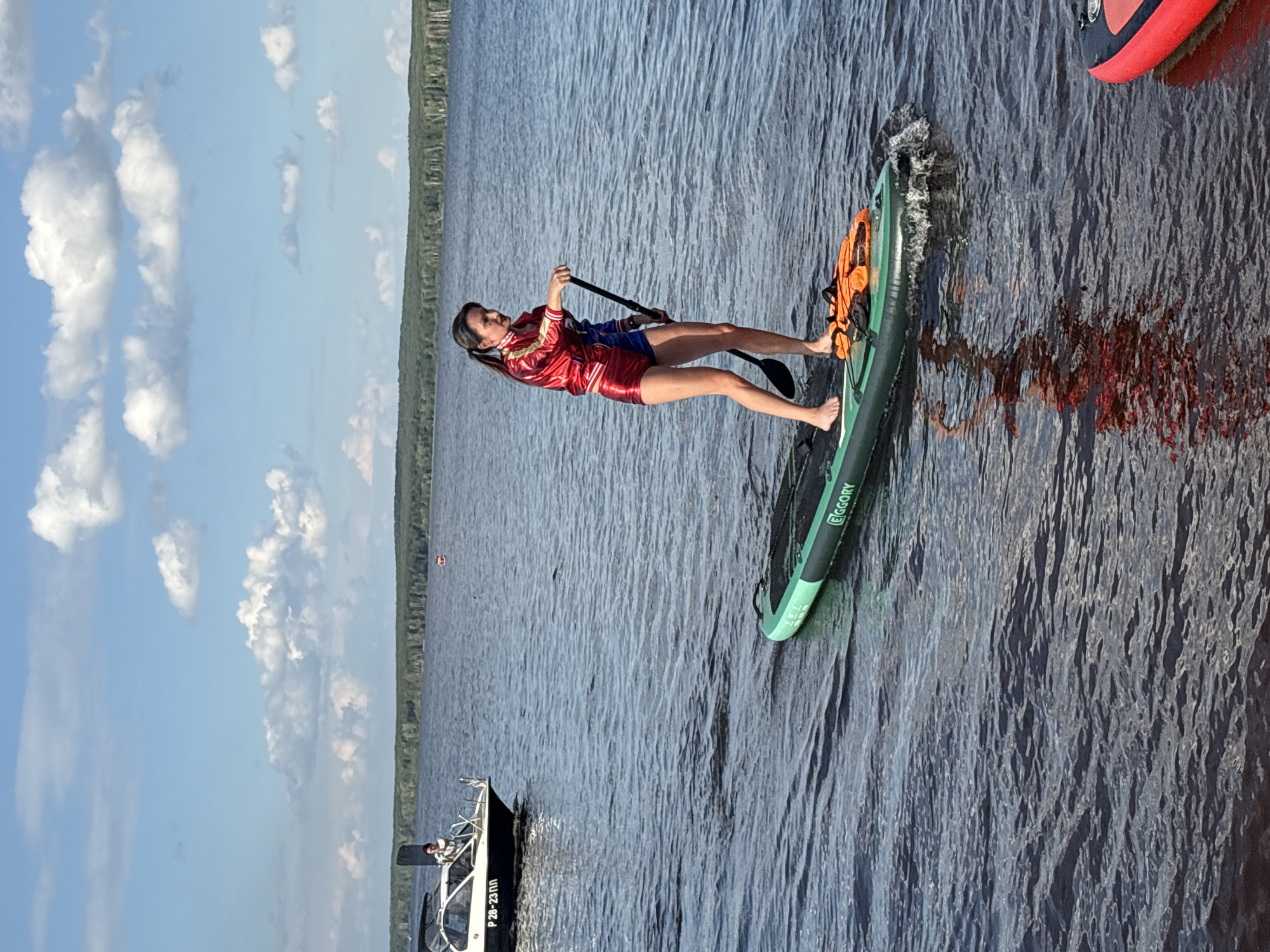 A woman in a red and blue costume resembling Harley Quinn is paddleboarding on a calm body of water. She is holding a paddle and looking to her left. The paddleboard is green and has an orange life vest on it. In the background, a white boat with a person standing on it is visible, and a tree line borders the distant shore under a blue sky with white clouds. The scene appears to be on the Kama River in the city of Chaikovsky, Russia, a popular recreational area.