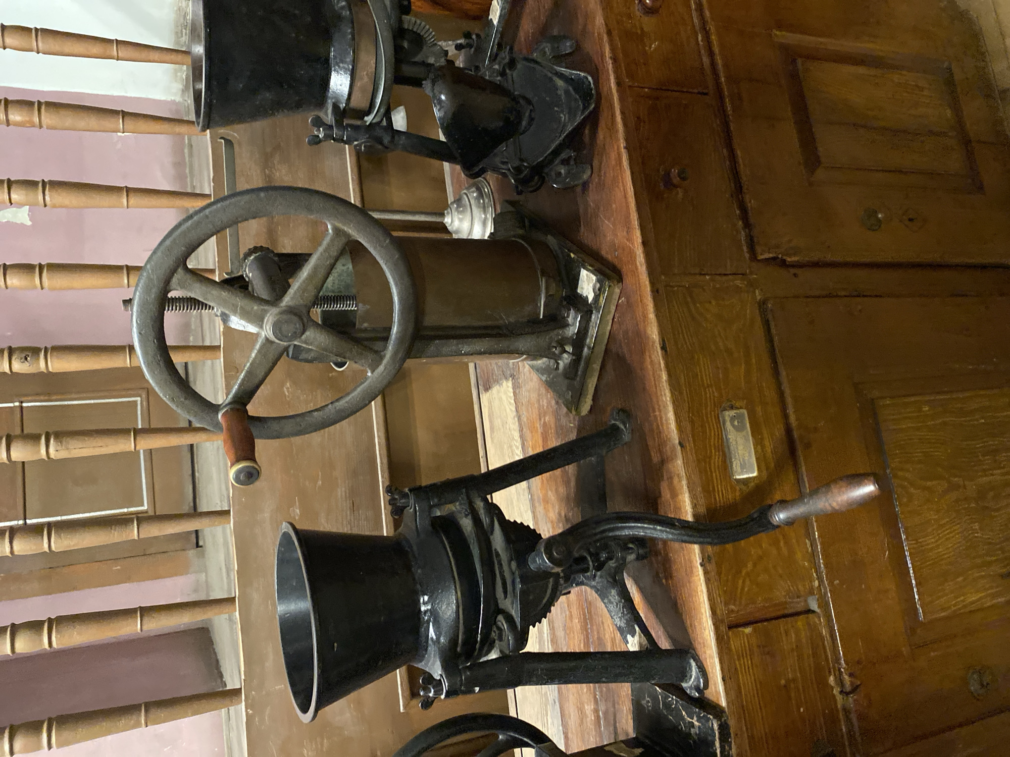 The image shows a close-up of vintage coffee grinders displayed on a wooden surface, likely a counter or cabinet. There are at least three grinders visible. The grinder on the left is black cast iron with a wide hopper, a mechanism below, and a hand crank with a wooden handle extending downwards. To its right is a taller grinder with a large, multi-spoked metal wheel for operation, a cylindrical body, and a crank mechanism. Further to the right is another black cast iron grinder, similar in style to the first, also featuring a hopper and visible working parts. The grinders are positioned on a wooden surface that appears to be part of an old cabinet with drawers and paneled doors. In the background, there are vertical wooden spindles, suggesting a railing or part of a piece of furniture. The lighting is ambient, highlighting the textures and aged appearance of the objects. The scene is devoid of people. This collection of antique coffee grinders is part of the exhibit at the Lviv Coffee Mining Manufacture in Lviv, Ukraine, which showcases the history and process of coffee.