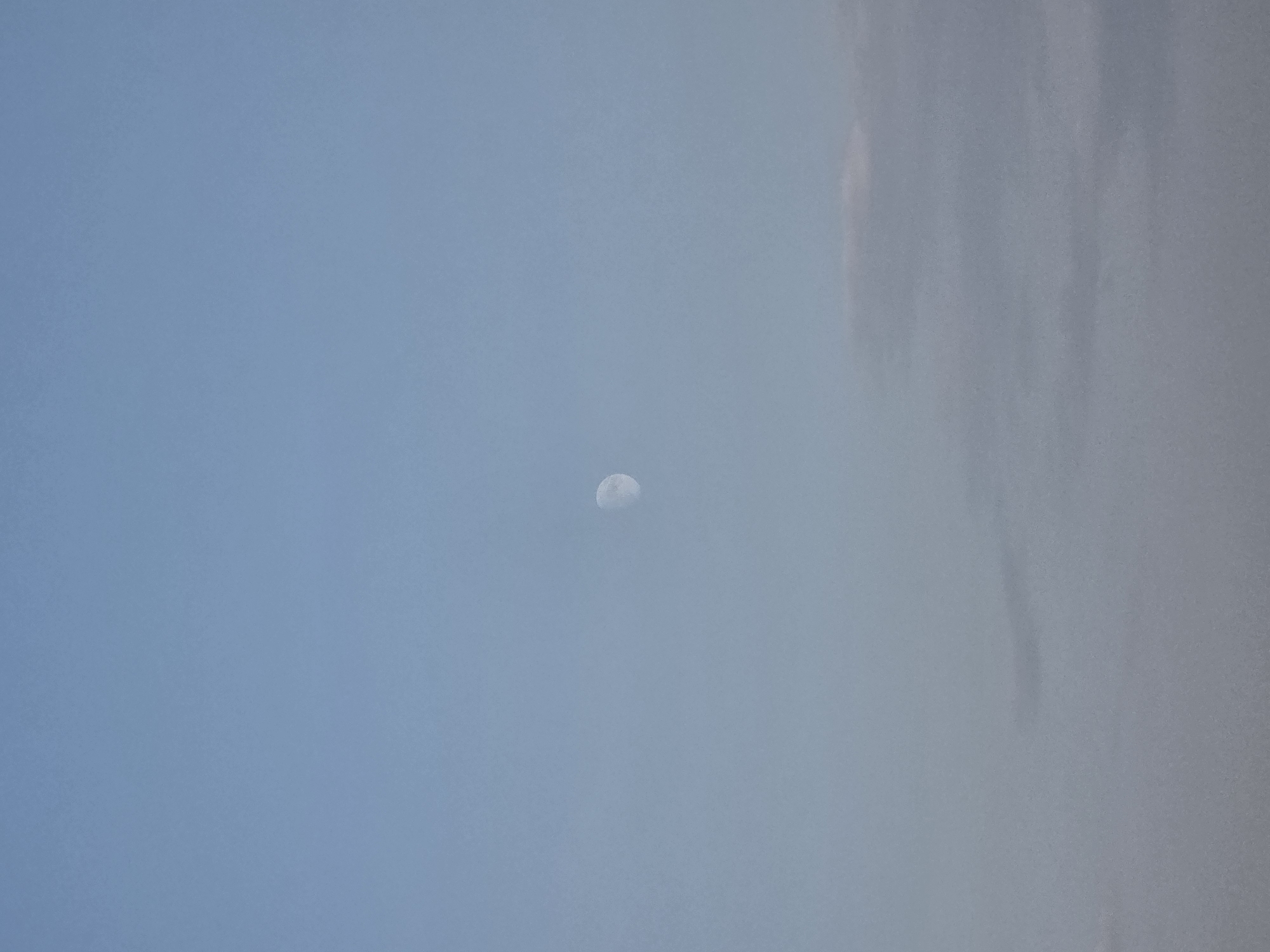 The image shows a hazy sky with a crescent moon visible in the upper center. The lower portion of the image contains indistinct clouds. There are no people, structures, or any other discernible objects or activities visible. The scene appears to be an outdoor environment, likely during daylight or twilight, given the presence of the moon and the clear sky, though the haze obscures detail. The location is identified as Obrochishte, Bulgaria, though the image itself does not contain any elements that specifically indicate this location.