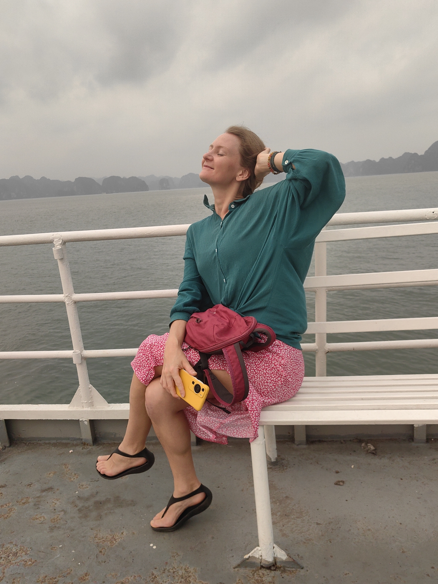 A woman sits on a white bench on a boat. She wears a teal long-sleeved shirt, a pink patterned skirt, and black sandals. She is holding a yellow smartphone in her right hand and has a red fanny pack on her lap. Her eyes are closed, her head is tilted back, and her left hand is behind her head. The background shows the sea and distant islands under a cloudy sky. The boat has a white railing. The location is Tuan Chau Ward, Vietnam.