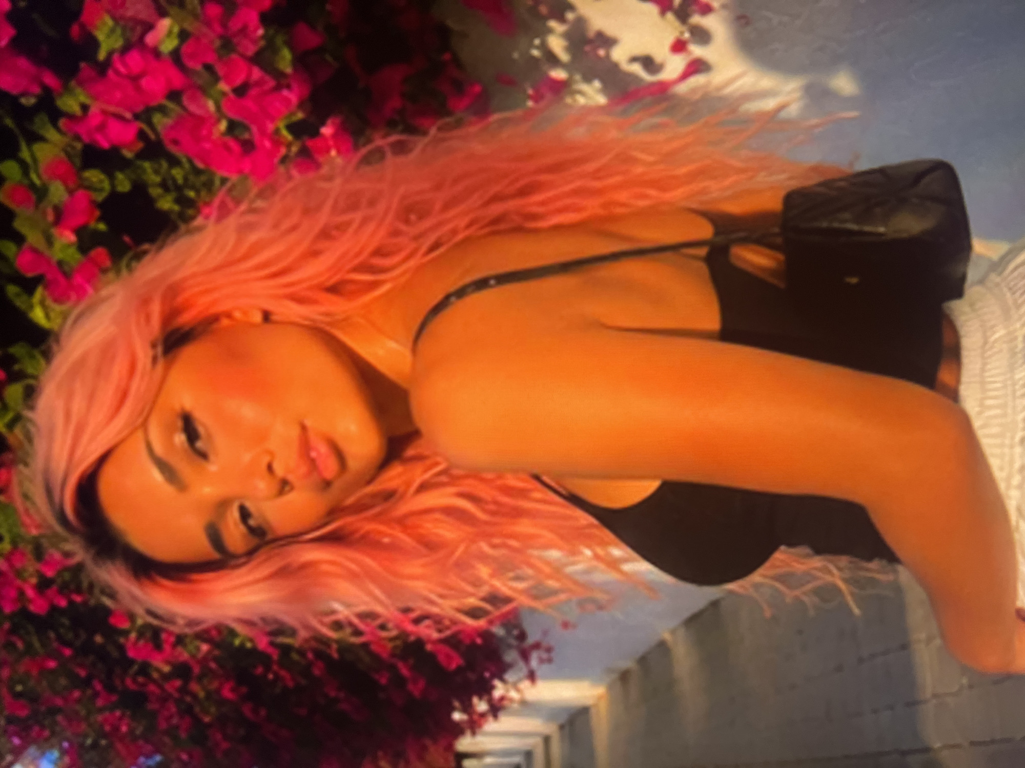 A woman with long, wavy, pink hair stands outdoors, looking back over her shoulder. She is wearing a black tank top and white pants. A small, black, quilted purse is slung over her shoulder. Behind her, vibrant pink flowers bloom on a bush, and a paved walkway runs alongside a white wall. The lighting suggests it is either sunrise or sunset, casting a warm, golden glow on the scene.