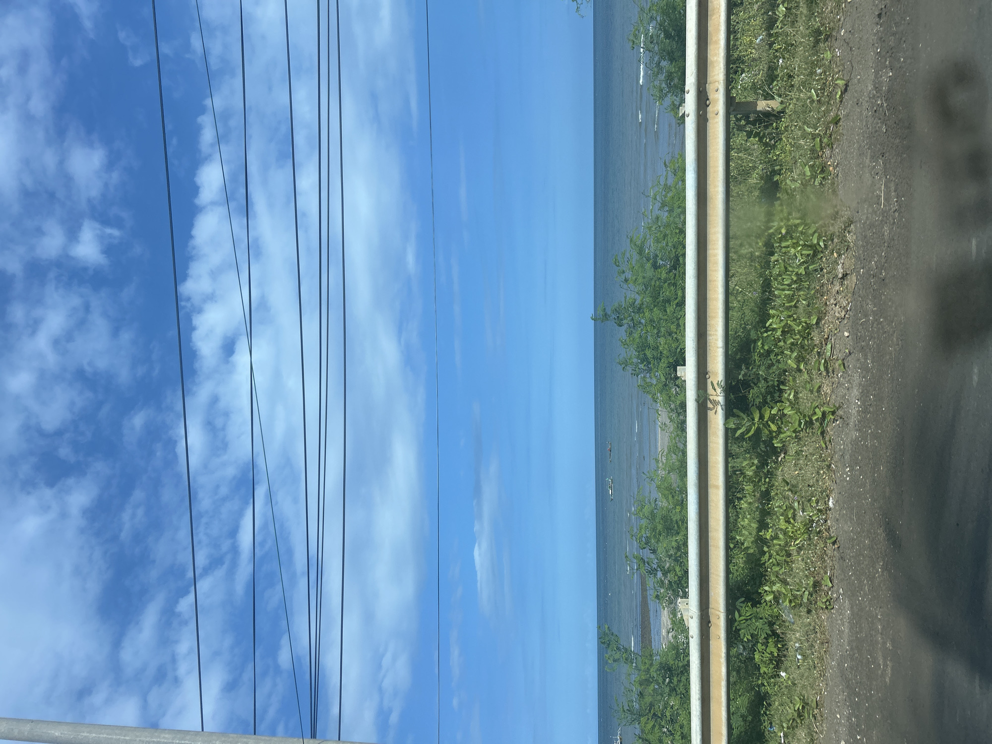 The image depicts a coastal scene with a clear blue sky populated by scattered white clouds. Several power lines are strung across the sky, supported by a vertical pole visible on the left side of the frame. In the middle ground, the ocean stretches out, with a few small boats visible on the water. A line of green vegetation, including bushes and small trees, runs along the shoreline. In the foreground, a weathered yellow guardrail separates the vegetation from a dark asphalt surface, likely a road. The environment appears to be dry with sparse green grass and some scattered debris near the guardrail. This scene is located in Borongloe, Indonesia.