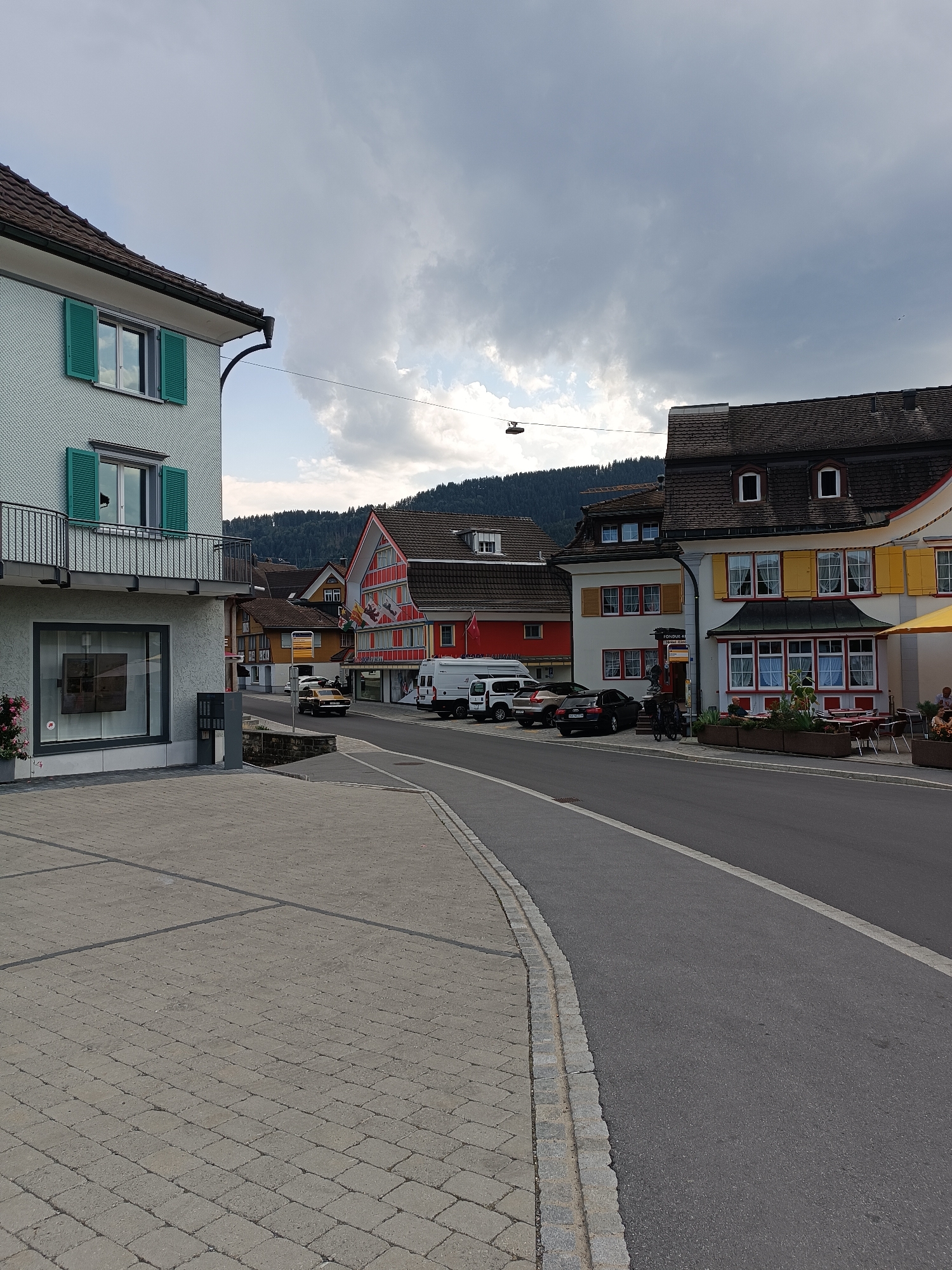 The image depicts a street scene in the town of Appenzell, Switzerland. On the left, a pale blue building with green shutters and a balcony is visible. A paved walkway with a light grey textured surface leads away from the building. The street itself is paved with asphalt and has a white lane marking.
Across the street, several buildings line the roadway. One building has a red and white facade with a steep, dark roof. Another building is painted yellow with red trim and features yellow shutters. Several cars are parked along the street, including a dark sedan, a silver car, and a gold-colored vintage car. A white van is also present.
In the background, a forested hillside rises under a cloudy sky. The overall atmosphere is that of a quiet, European town.