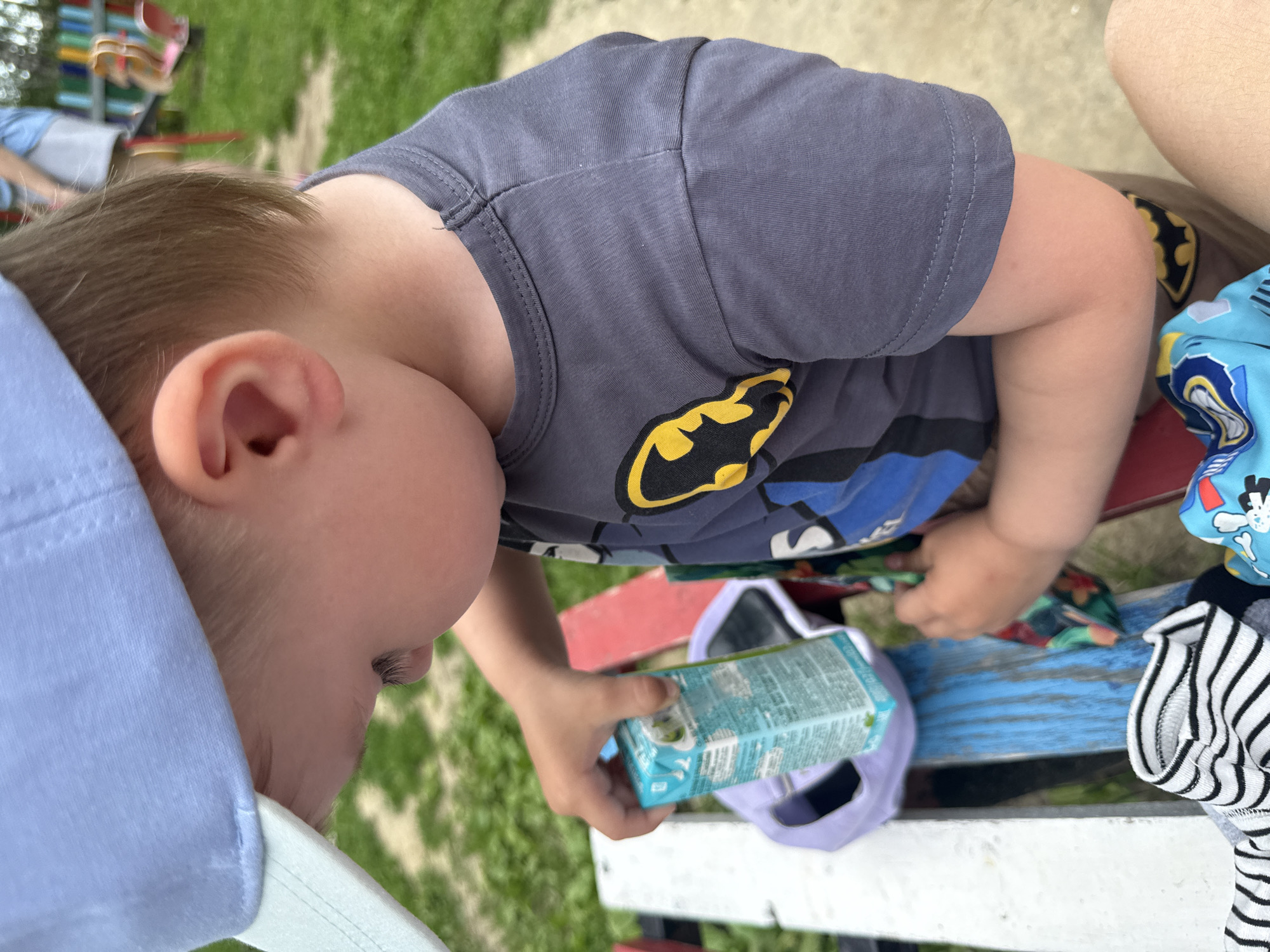The image shows a close-up of a young child wearing a light blue baseball cap and a grey t-shirt with a Batman logo. The child is holding a small carton of juice. They are seated on a bench with red and blue painted slats. In the background, a playground can be seen with colorful equipment, suggesting the location is a park or playground in the city of Oktyabrskoye, Russia. The child appears to be engrossed in examining the juice carton. The focus is on the child and their immediate surroundings, with the background slightly blurred.