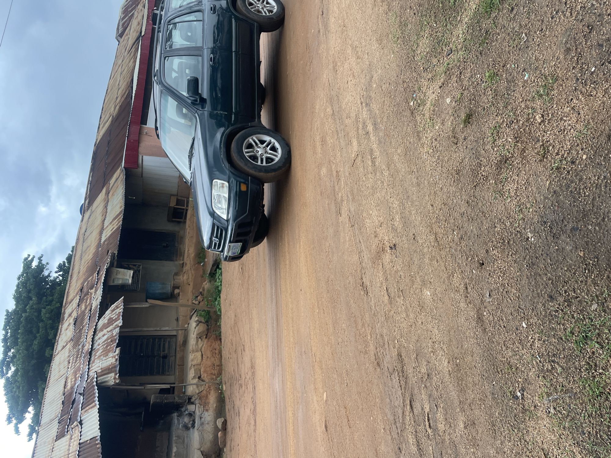 A dark green Honda CR-V is parked on a dirt road in front of a building with a rusted corrugated metal roof. The building appears to be a shop or dwelling, with a rough concrete foundation and what looks like wooden doors and shutters. There is evidence of previous construction or repairs on the building, with various materials visible. The ground is a reddish-brown dirt, with some patches of dry grass and scattered stones. The sky is overcast with a light gray cloud cover, suggesting a potentially humid or rainy day. The overall impression is a rural or semi-urban setting in Gwong, Nigeria, characterized by simple architecture and unpaved roads. There are no people visible in the image.
