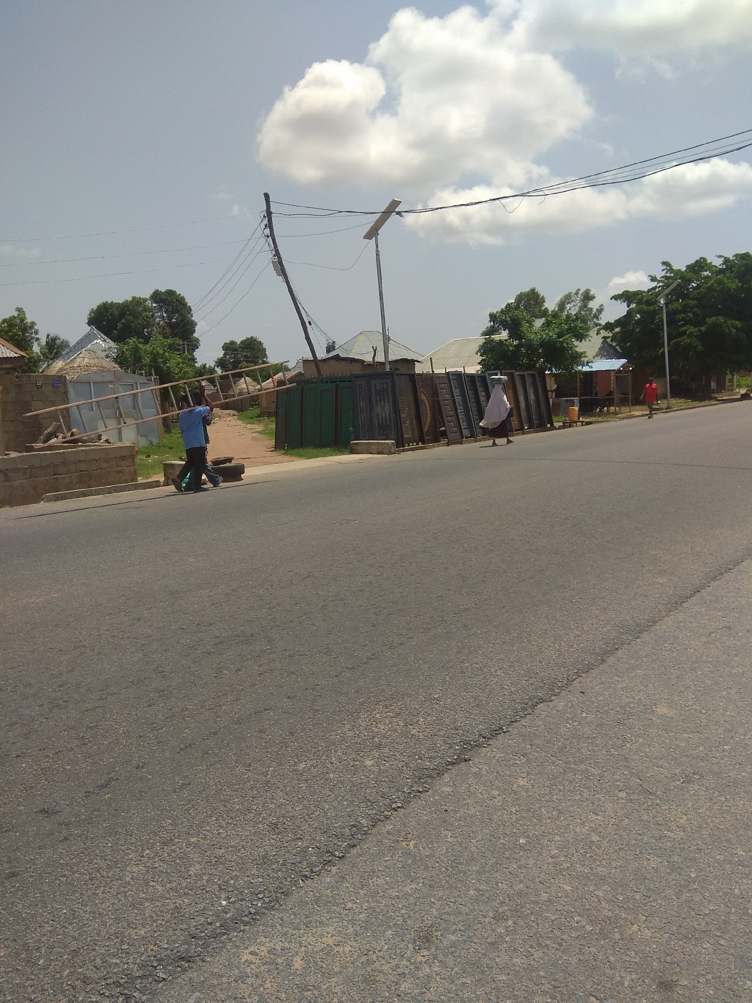 The image depicts a street scene in Jalingo, Nigeria, on a bright, cloudy day. In the foreground, an asphalt road stretches into the distance. On the left side of the road, two men are walking away from the camera, carrying a long wooden ladder. They are walking on the sidewalk, which is bordered by a low brick wall. Behind the men and the wall, there are several structures, including buildings with corrugated metal roofs and some thatched roofs.
Further down the street, to the right of the men with the ladder, a display of metal gates or panels stands upright along the roadside. A person wearing a white headscarf and dark clothing is walking past these gates. Another person in a red shirt is walking further down the street on the right side of the image.
The environment includes trees and vegetation scattered throughout the background. Overhead, power lines are visible, supported by poles, and a solar-powered street light is mounted on a pole. The sky is partially covered with white, puffy clouds. The overall impression is a view of a moderately busy street in a town or city in Nigeria.