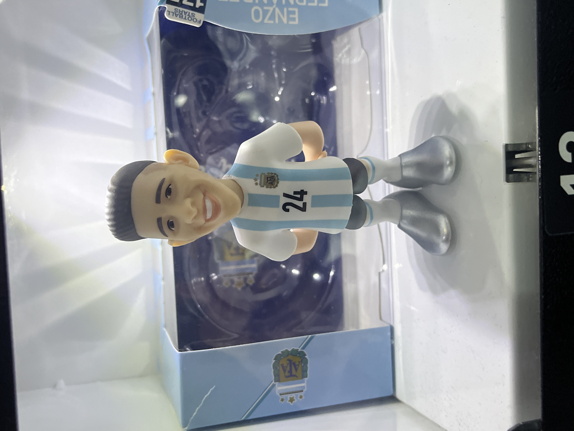 An indoor, brightly lit, close-up image displays a collectible figurine of a male football player, presented within a clear plastic and cardboard packaging box. The image itself is rotated 90 degrees counter-clockwise, making the figurine appear to lie horizontally.
The figurine depicts a young man with dark hair, a smiling expression, and fair skin. He is dressed in a light blue and white striped football jersey, typical of the Argentina national team, with dark shorts and metallic silver boots. The number "24" is prominently displayed on the front of his jersey, along with the Argentinian Football Association (AFA) logo, featuring three stars above the "AFA" lettering.
The figurine is encased in a clear plastic blister pack, which is mounted on a light blue cardboard backing. The top section of the packaging, visible at the image's upper edge, features text that appears inverted due to the image's rotation; it reads "ENZO FERNANDEZ" and "FOOTBALL STARS," with "17S" also partially visible. The bottom section of the cardboard packaging also displays the AFA logo with three stars.
The figurine and its packaging are set against a bright, largely white background, indicating an indoor environment. A dark, possibly black, vertical structure with a metal bracket is visible along the right edge of the frame, suggesting the item is on display, perhaps in a store, a cabinet, or a vending machine. While the context of Ulyanovsk, Russia, is provided, there are no specific visual cues within the image to confirm this location.