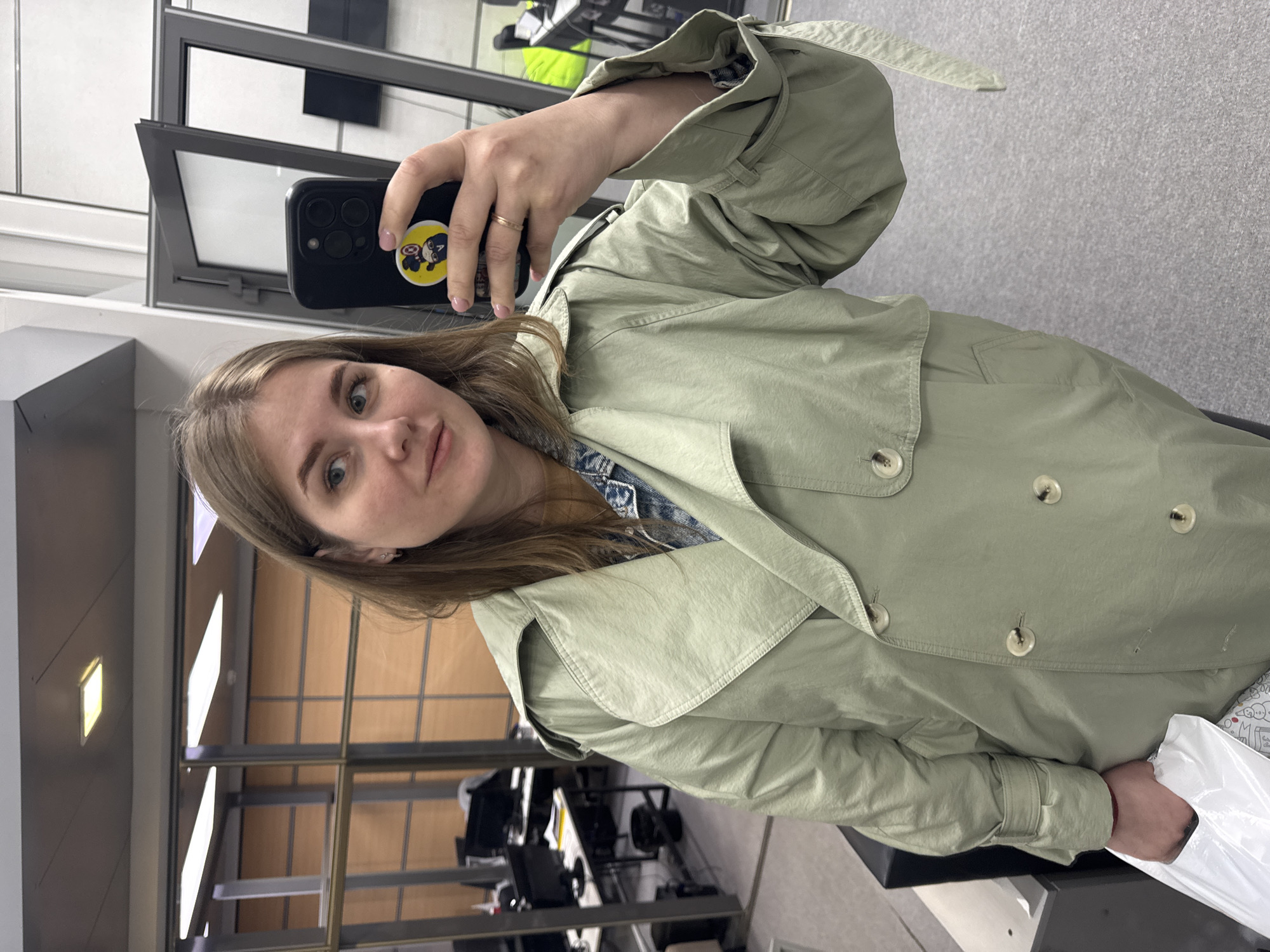 A woman is taking a selfie in a building in Perm, Russia. She has shoulder-length brown hair and is wearing a light green trench coat over a denim jacket. She is holding a black smartphone in her right hand, which has a sticker of Captain America on the back. The phone's screen is obscured. She is wearing a ring on her ring finger.
The background includes a glass wall with vertical supports and a gray floor. In the background, office chairs and desks are visible, along with parts of the building's interior including walls and lighting fixtures. The overall lighting is bright, suggesting an indoor location during daylight hours. She is also holding a white plastic bag in her left hand.
