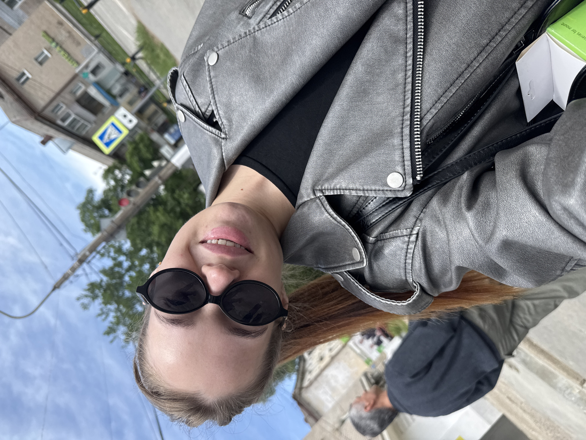 The image shows a woman wearing black sunglasses and a silver jacket. She appears to be outdoors in Chaikovsky, Russia, as indicated by the presence of a "Pedestrian Crossing" sign typical of the city. Behind her, a man with gray hair and a dark jacket is partially visible. The background includes a building and trees, suggesting an urban environment.