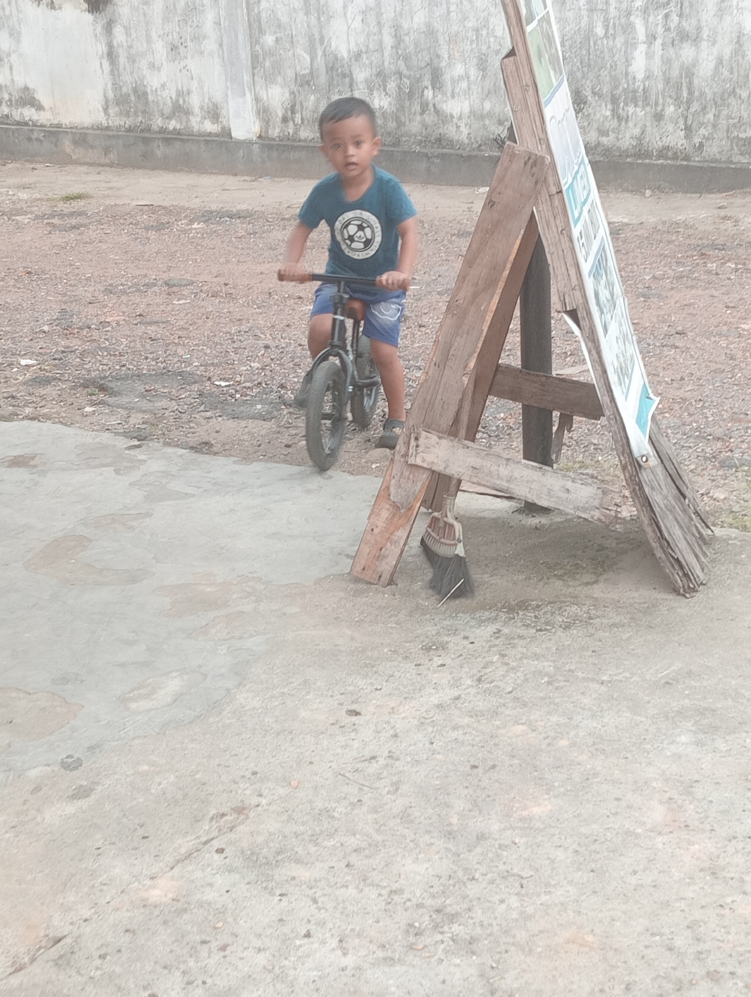 In Jambi, Indonesia, a young boy rides a small bicycle on a gravel surface. He is wearing a dark blue t-shirt with a circular emblem and blue shorts. The bicycle has black tires and handlebars. Behind him, a weathered wooden A-frame sign stands, displaying various posters. A broom is propped against the sign. The background features a textured wall and the ground transitions from gravel to concrete.