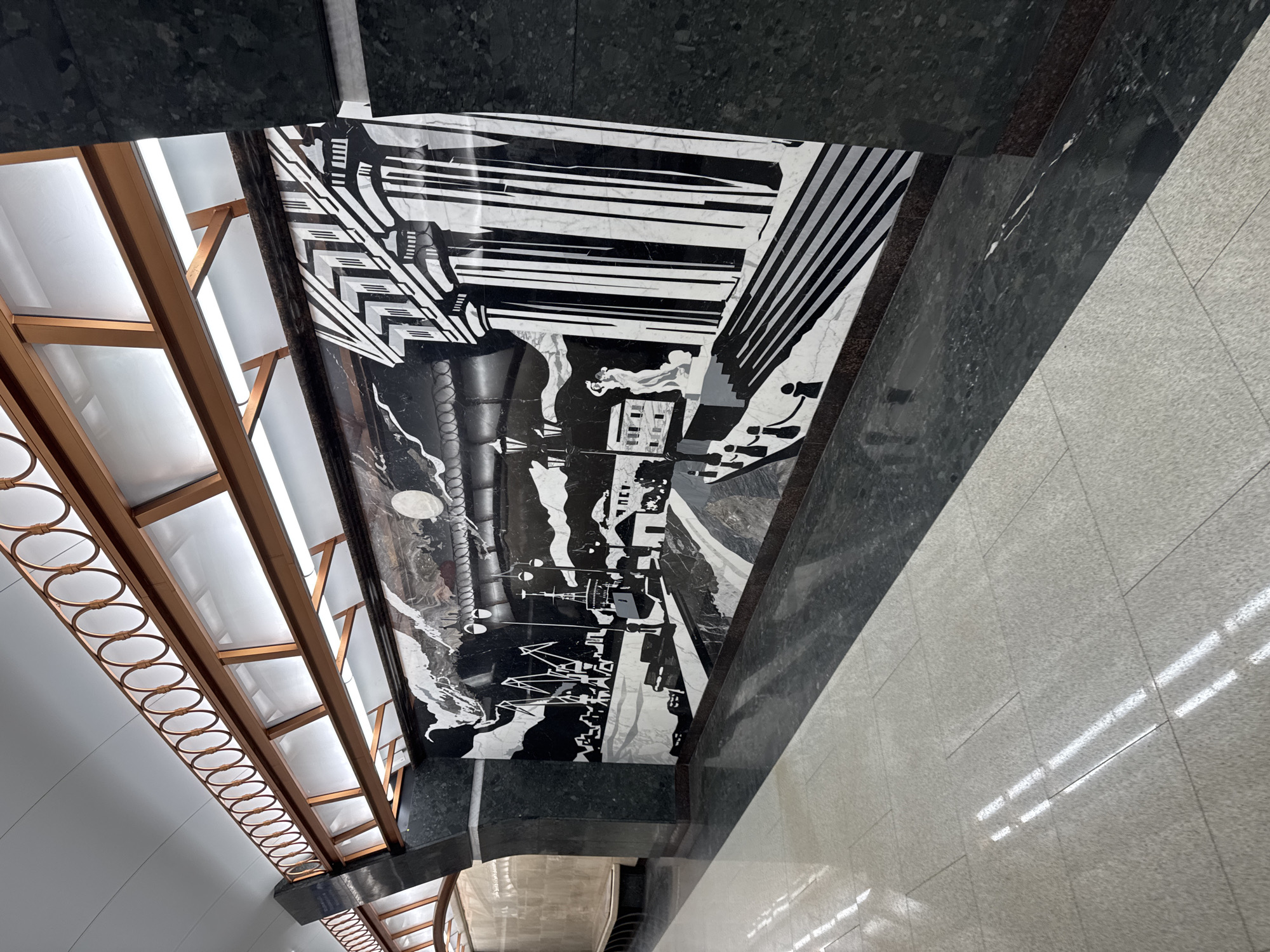 The image displays a grand architectural interior, most likely a metro station in Saint Petersburg, Russia. The dominant feature is a large, black and white marble inlay mural or mosaic covering a substantial section of what appears to be an arched ceiling or high wall. The mural depicts a stylized, geometric cityscape in a graphic, high-contrast style reminiscent of Constructivist or Art Deco art. It includes abstract buildings, industrial forms, and what appears to be a large, circular moon or sun disc, with hints of figures integrated into the landscape.
Adjacent to the mural, a decorative lighting fixture runs along the length of the space. This fixture comprises a golden or bronze-colored metallic framework, featuring a repeating pattern of interconnected circular rings. Behind this framework, translucent white panels diffuse light from visible fluorescent tubes, providing indirect illumination.
The mural and lighting system are flanked by dark, highly polished granite or marble surfaces on the surrounding walls. The floor is made of light-colored, possibly grey, tiled material, reflecting the bright overhead lights. The overall aesthetic is monumental, clean, and meticulously designed, reflecting the ornate and artistic characteristics of public spaces from the Soviet era common in the Saint Petersburg metro. No people are visible, and there is no discernible text in the image. The environment is brightly lit, suggesting a well-maintained interior.