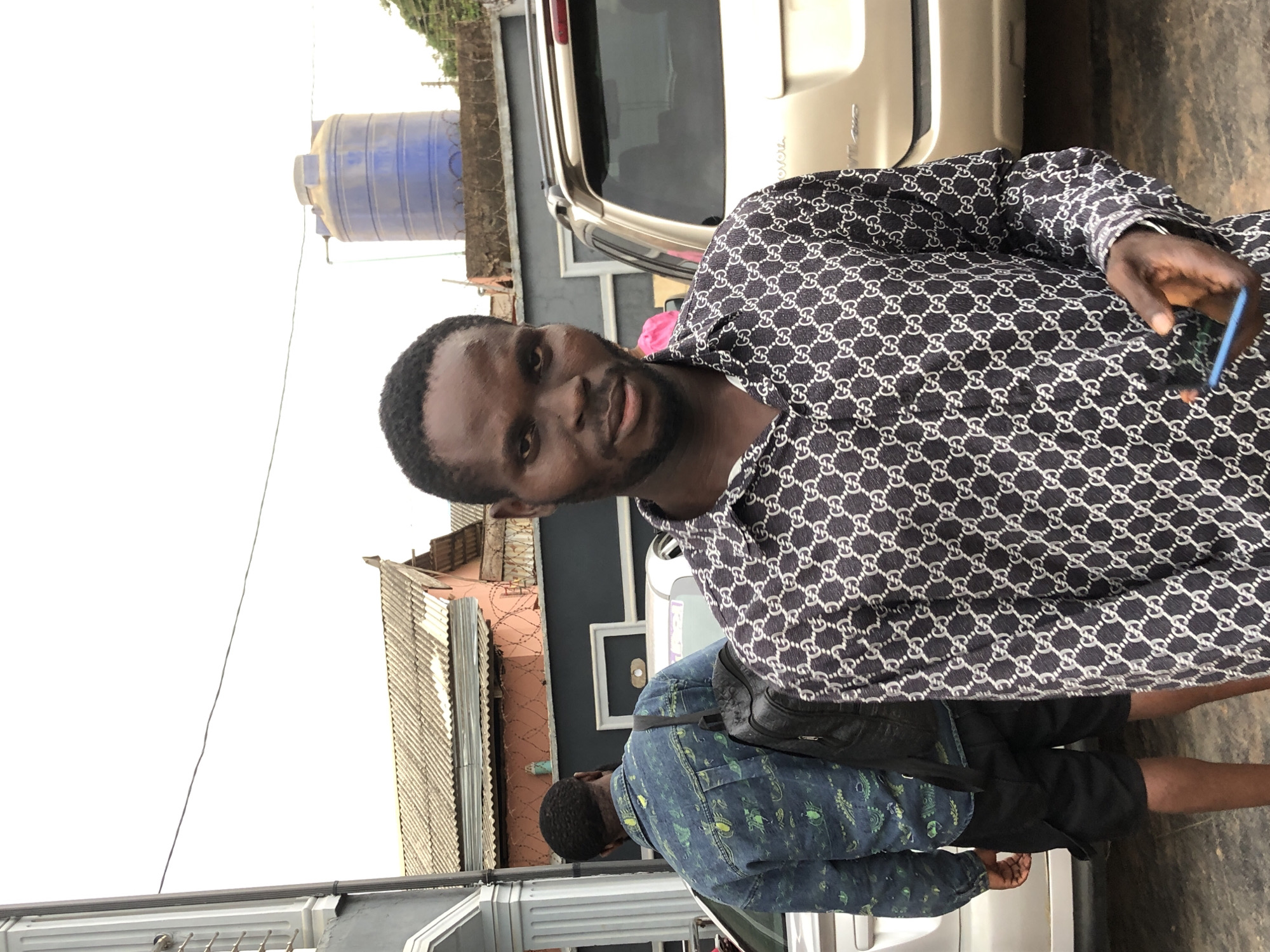 An outdoor, eye-level shot captures a man posing for the camera in an urban environment during daylight. The primary subject, a man with dark skin, short dark hair, and a short beard, occupies the left side of the frame, looking directly at the viewer with a neutral to slightly smiling expression. He is wearing a long-sleeved dark blue or black shirt with a repeating white "GG" pattern, reminiscent of the Gucci monogram. He holds a light blue smartphone in his left hand.
Behind him, on the right side of the image, another man is partially visible from the back, appearing to walk away. He wears a blue patterned shirt or jacket, dark shorts, and a black shoulder bag. In the background, a light-colored SUV with "Toyota" visible on its rear is parked. Further back, a large blue plastic water tank sits on a stand. Residential or commercial buildings with corrugated metal roofs and walls of varying colors (pink, dark blue) are visible. Power lines or wires stretch across the bright, almost white sky. The setting suggests a street or a developed area. The bright sky indicates either a clear day or an overcast one.