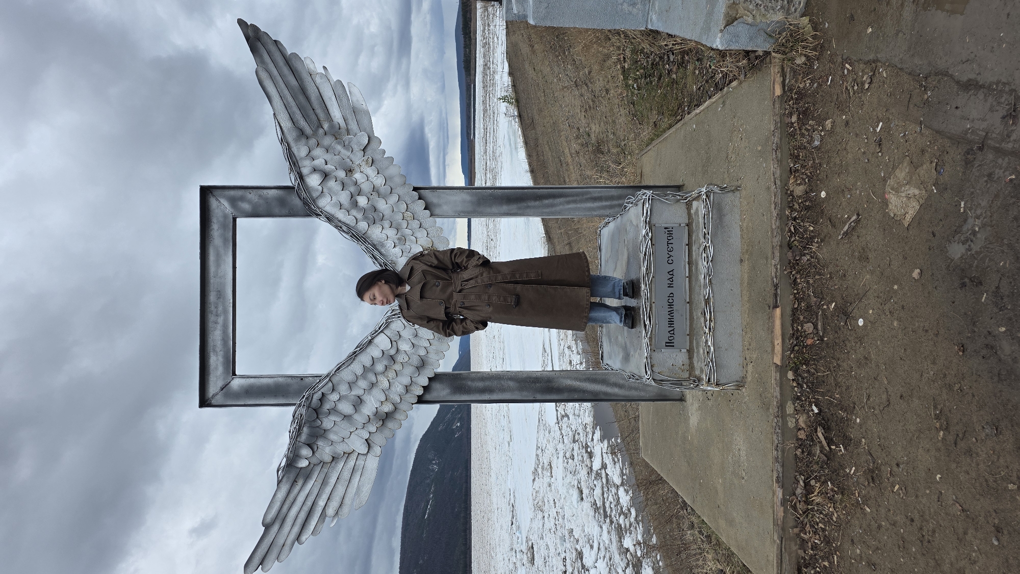 A woman in a brown trench coat stands within an angel wings sculpture, positioned outdoors in Lensk, Russia. The sculpture consists of a square frame with two large angel wings attached to its sides. The base of the structure, adorned with metal chains, bears an inscription in Cyrillic script that reads "Поднимись над суетой!", which translates to "Rise above the bustle!". The surrounding environment includes a landscape with a body of water, visible ice floes, distant mountains, and a cloudy sky. The ground near the sculpture is a mix of dirt and concrete.