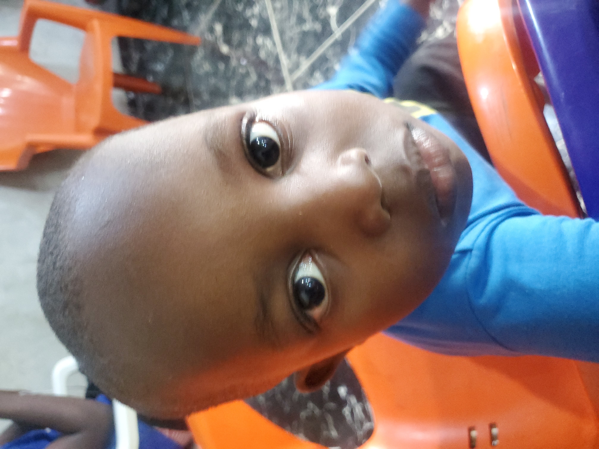 The image shows a close-up of a young child's face, looking directly at the camera. The child has dark skin and short, dark hair. They are wearing a bright blue long-sleeved shirt. The child is seated in an orange chair, and a portion of a blue object is visible in the foreground, possibly part of a toy or another chair. In the background, another orange chair is visible, as well as what appears to be a tiled floor. The lighting suggests an indoor environment. No specific location in the city of Unknown, Unknown can be determined.
