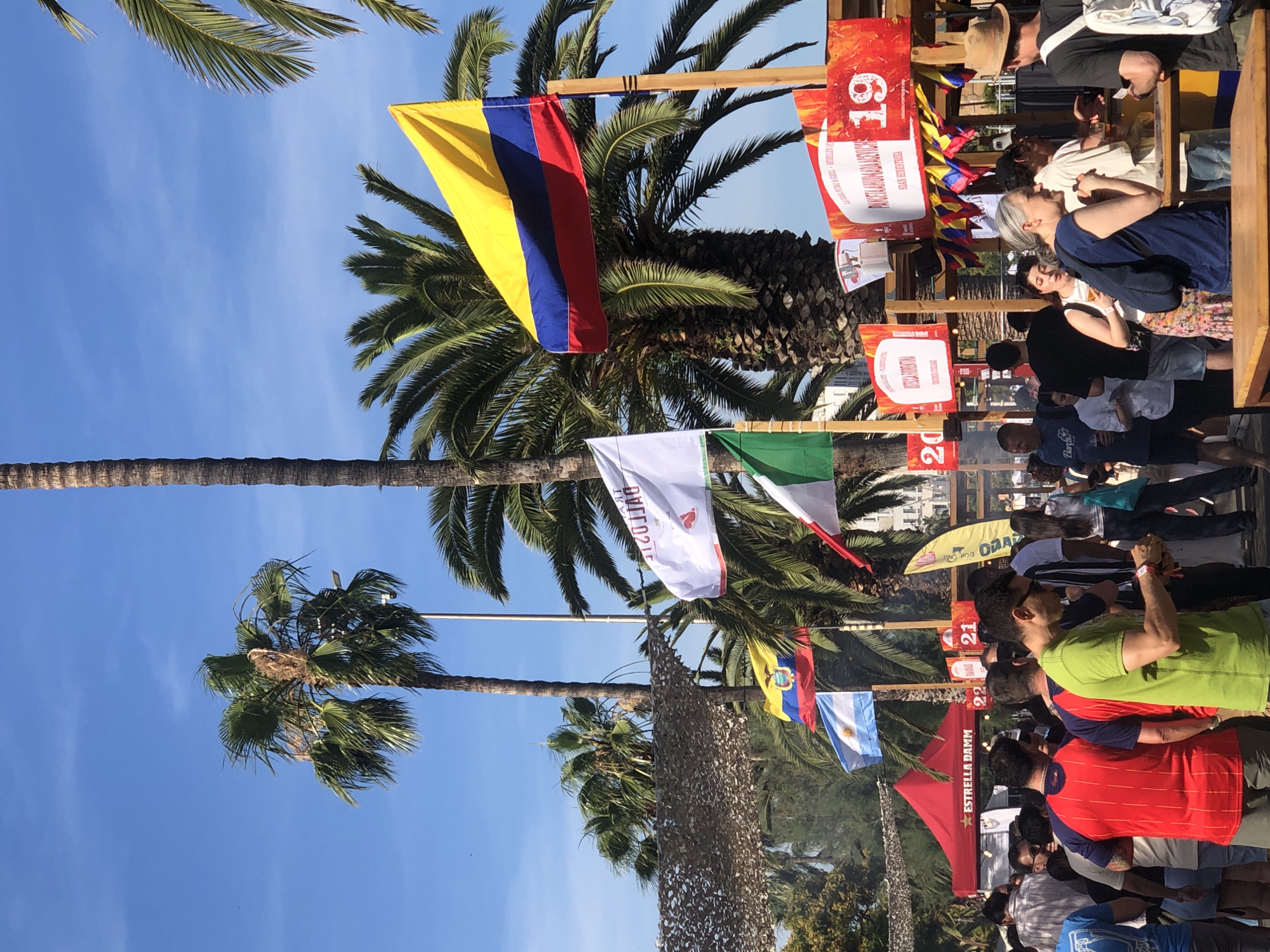 The image depicts an outdoor market or festival scene, likely in a warm climate given the palm trees and bright sunlight. Several flags are visible, including the Colombian flag, the Ecuadorian flag, and the Argentinian flag, suggesting a cultural event or food festival with an international theme. Numerous people are present, browsing stalls, interacting, and in motion. There are several food stalls with signage, some of which display numbers like "19" and "20," possibly indicating stall numbers or menu items. One stall has a prominent red tent with the "Estrella Damm" logo. The environment includes palm trees, a clear blue sky, and some overhead netting.