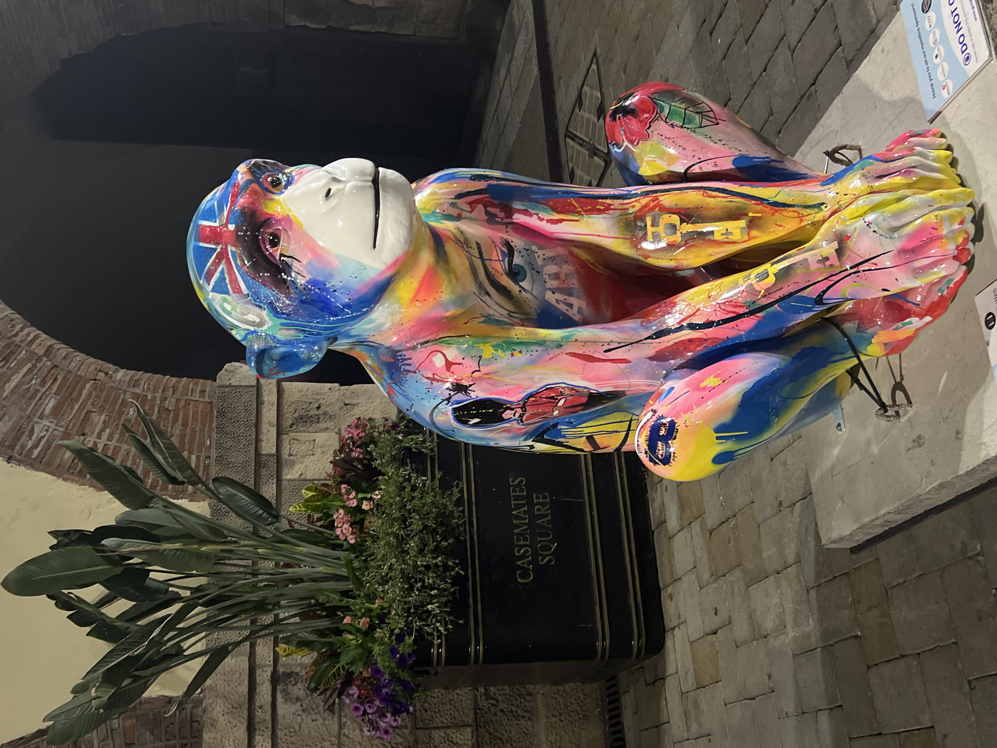 This is a medium shot, eye-level, outdoor, nighttime photograph of a brightly colored, art installation monkey statue in Casemates Square, Gibraltar. The monkey is painted with a variety of vibrant colors and patterns, including splashes of pink, yellow, blue, and green, as well as images of a British flag on its head, an eye on its torso, and a military figure on its leg. The monkey is in a seated, crouched position, with its hands resting on its knees.
Behind the monkey, to the left, is a large, black planter filled with lush green plants and some smaller pink and purple flowers. The planter has "CASEMATES SQUARE" inscribed on its side in gold lettering. The background of the image shows a stone wall and an arched entryway, consistent with the architecture of Casemates Square. The ground is paved with large, rectangular stone tiles. A small sign with sponsor information is visible at the bottom right corner of the image. The lighting is dim, suggesting it is nighttime, and the colors of the monkey statue are highly saturated.