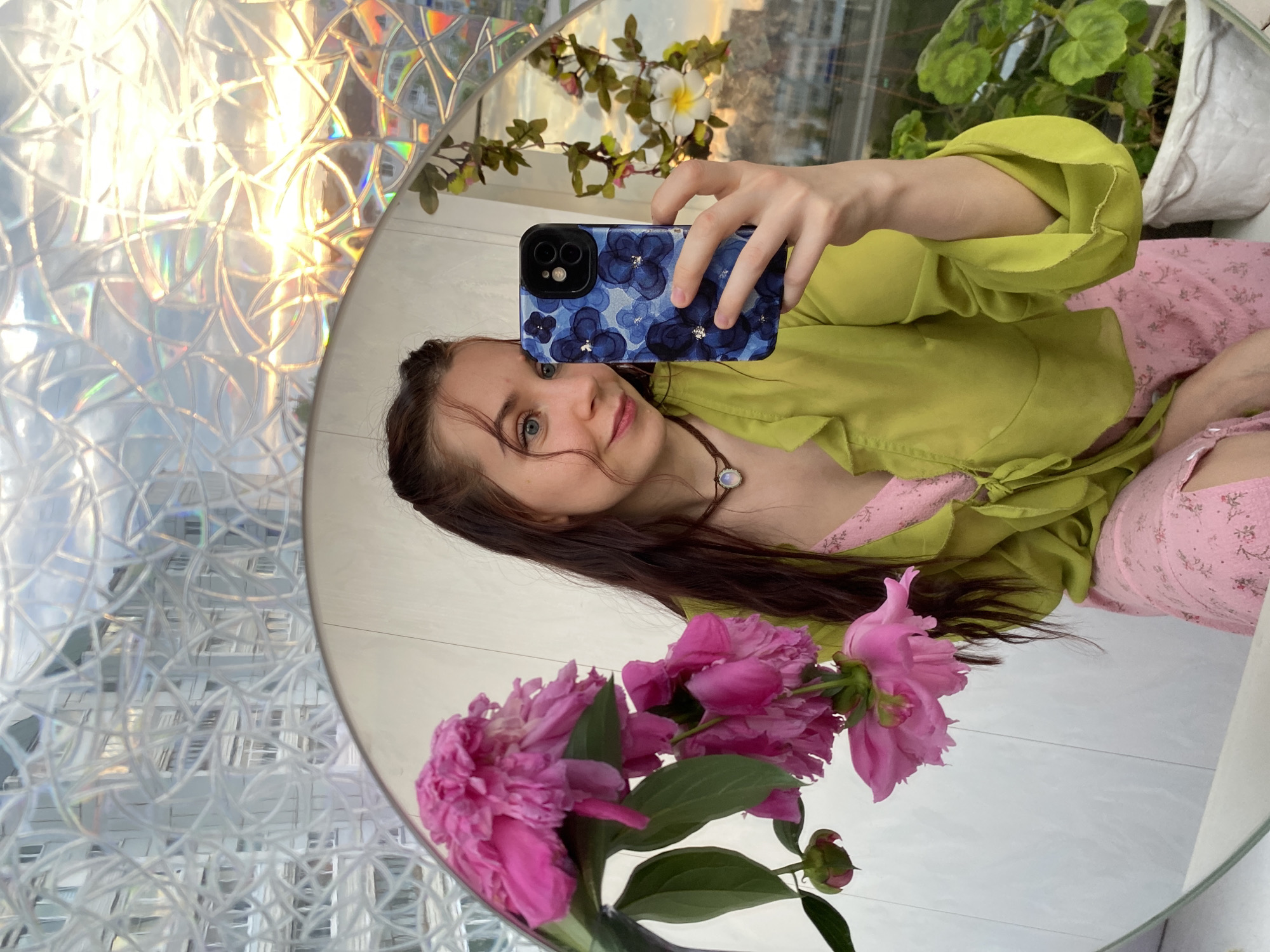 In a selfie taken in Chaikovsky, Russia, a young woman with long, dark hair stands before a mirror, holding up her phone to capture her reflection. She is wearing a pink floral dress and a lime-green, sheer blouse with ruffled sleeves. A bouquet of bright pink peonies is held to her left, partially obscuring the mirror's reflection. The background is a window with a textured, patterned film, through which the golden hues of a sunset can be seen illuminating buildings in the distance. Green plants in a white pot are visible on the windowsill to the right. The woman is smiling as she takes the photo.