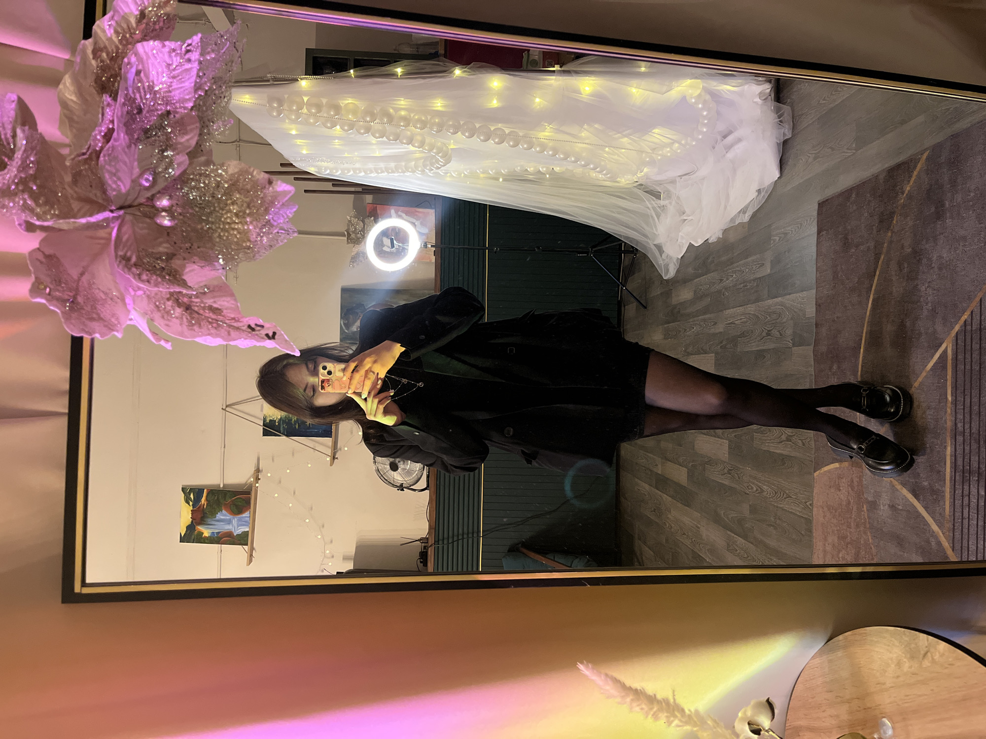 A young woman is taking a selfie in a mirror. She is wearing a black coat, black pantyhose, and black shoes. The mirror reflects a room with light hardwood floors and a dark gray rug. There is a backdrop with sheer white fabric and string lights. The mirror also reflects decorations, including a large pink and silver flower arrangement and a small tabletop. The location is likely a room in an apartment or studio. I don't have enough information to determine if this is in Saint Petersburg, Russia.