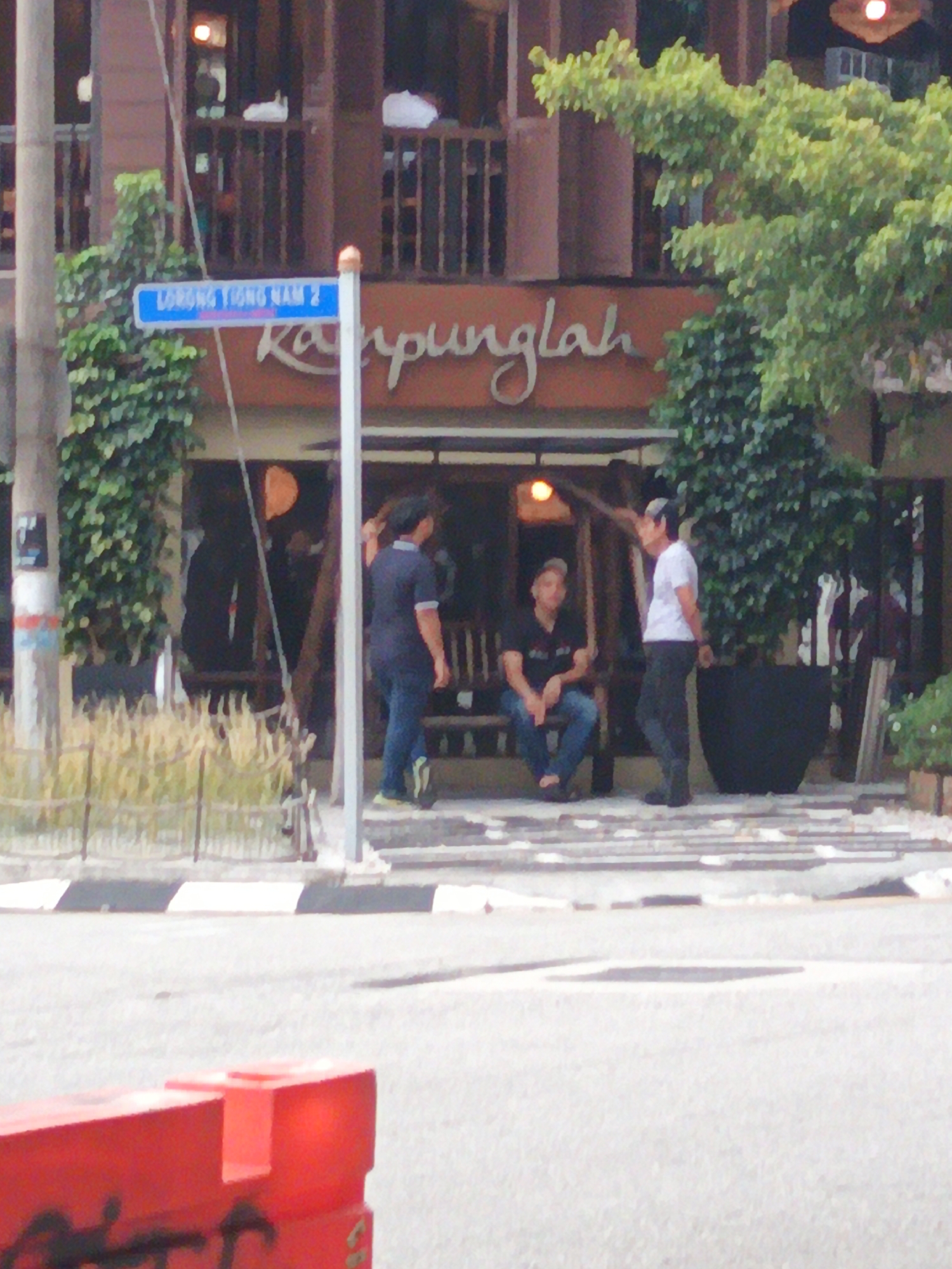 A street sign reading "LORONG TIONG NAM 2" is visible in the foreground of the image, suggesting the location is Tiong Nam Road, Kuala Lumpur, Malaysia. Behind the sign, a building with a brownish facade and wooden balcony railings is depicted. The building's name, "Kampunglah," is written in cursive script on its front. Three men are present outside the building. One man in a dark blue shirt and jeans stands to the left. A second man in a black shirt and jeans sits on a bench in the middle, while a third man in a white t-shirt and dark pants stands to the right. The scene is set on a street with a paved road and a sidewalk. Lush green trees and potted plants add to the environment. Red traffic barriers are also visible in the lower part of the image.