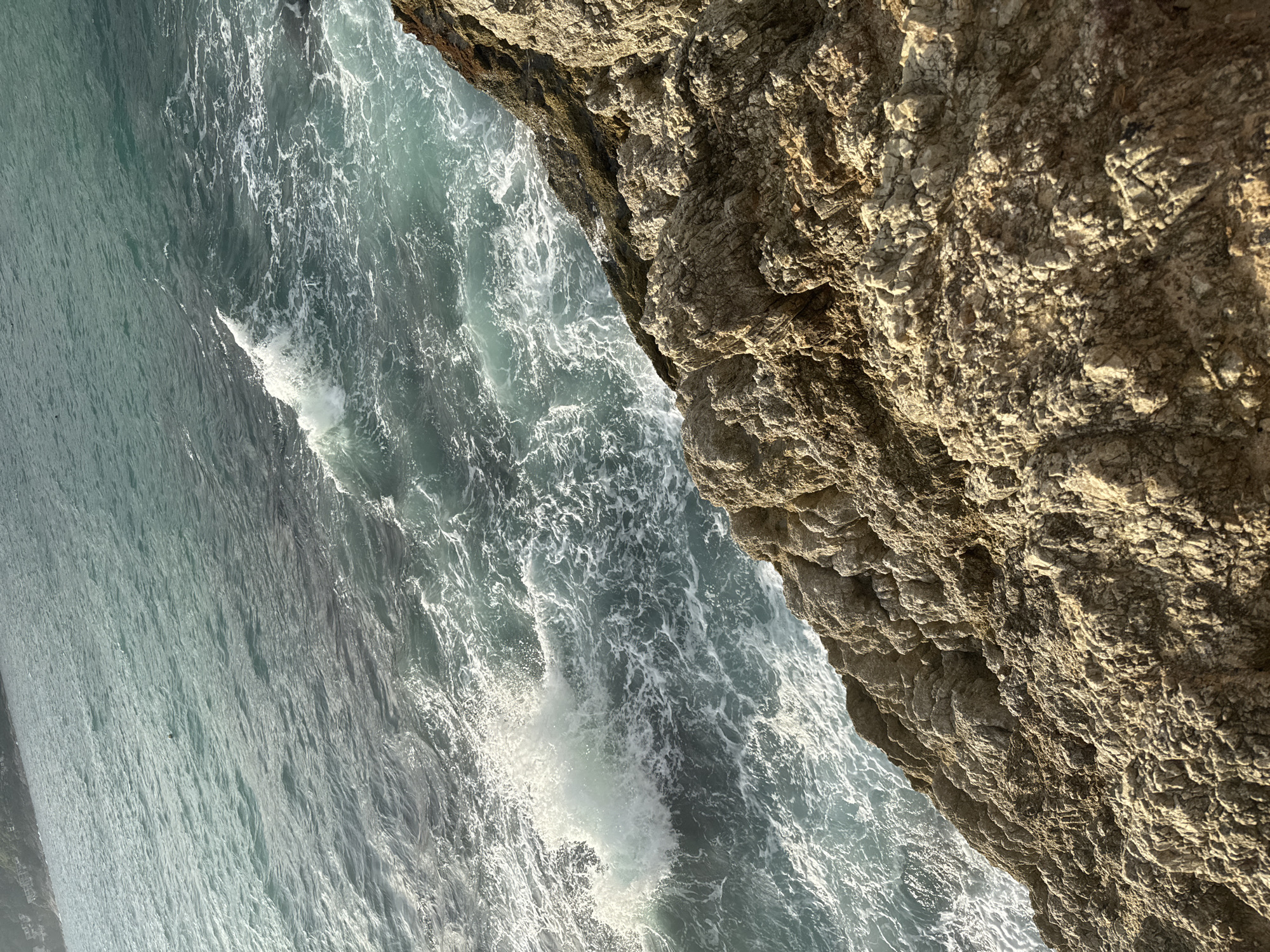 A high-angle medium shot captures the rugged, rocky coastline of Rawai, Thailand, where the turquoise ocean meets a steep cliff face. The water is dynamic, with waves crashing against the rocks, creating white foam and spray. The rocks themselves are textured and weathered, appearing to be a mixture of tan and gray. In the distance, the faint outline of buildings can be seen on a hillside, suggesting a developed area beyond the natural coastline. The lighting indicates it is daytime, likely late afternoon, as the sun casts shadows and highlights on the rocky terrain. No people or specific objects are clearly identifiable in the frame.