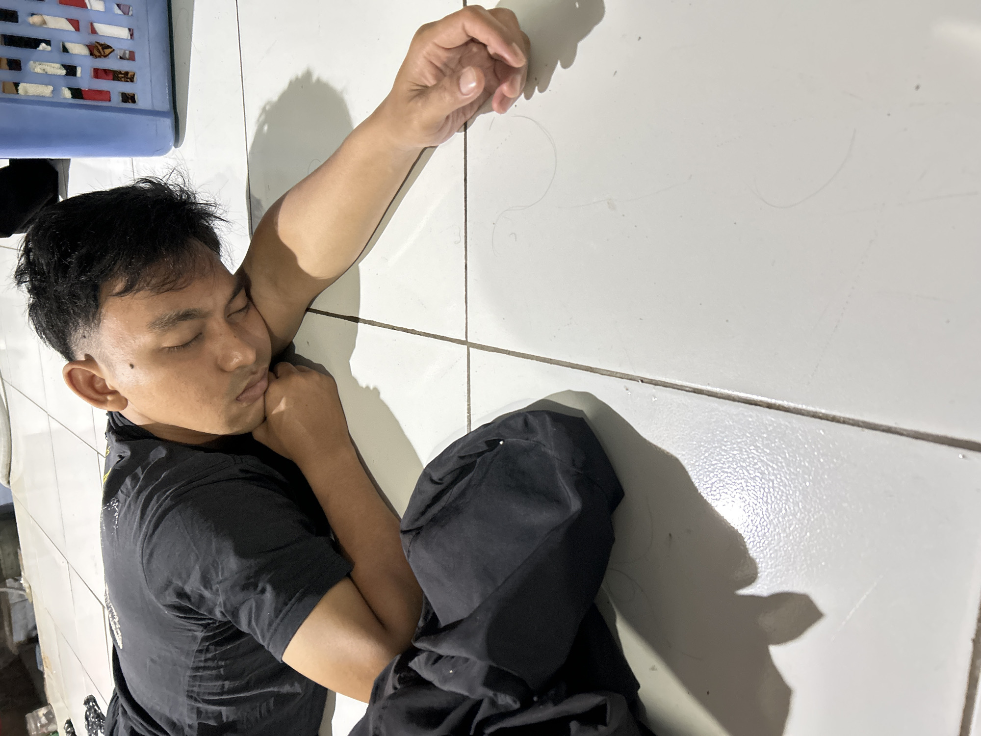 The image shows a man lying on the floor in what appears to be a residential setting in Depok, Indonesia. He is asleep, with his head resting on his arm. He is wearing a black t-shirt. A crumpled black garment is next to him. A blue plastic laundry basket filled with clothes is visible in the background. The floor is tiled. The lighting suggests it is indoors, possibly at night, with artificial light sources casting shadows.