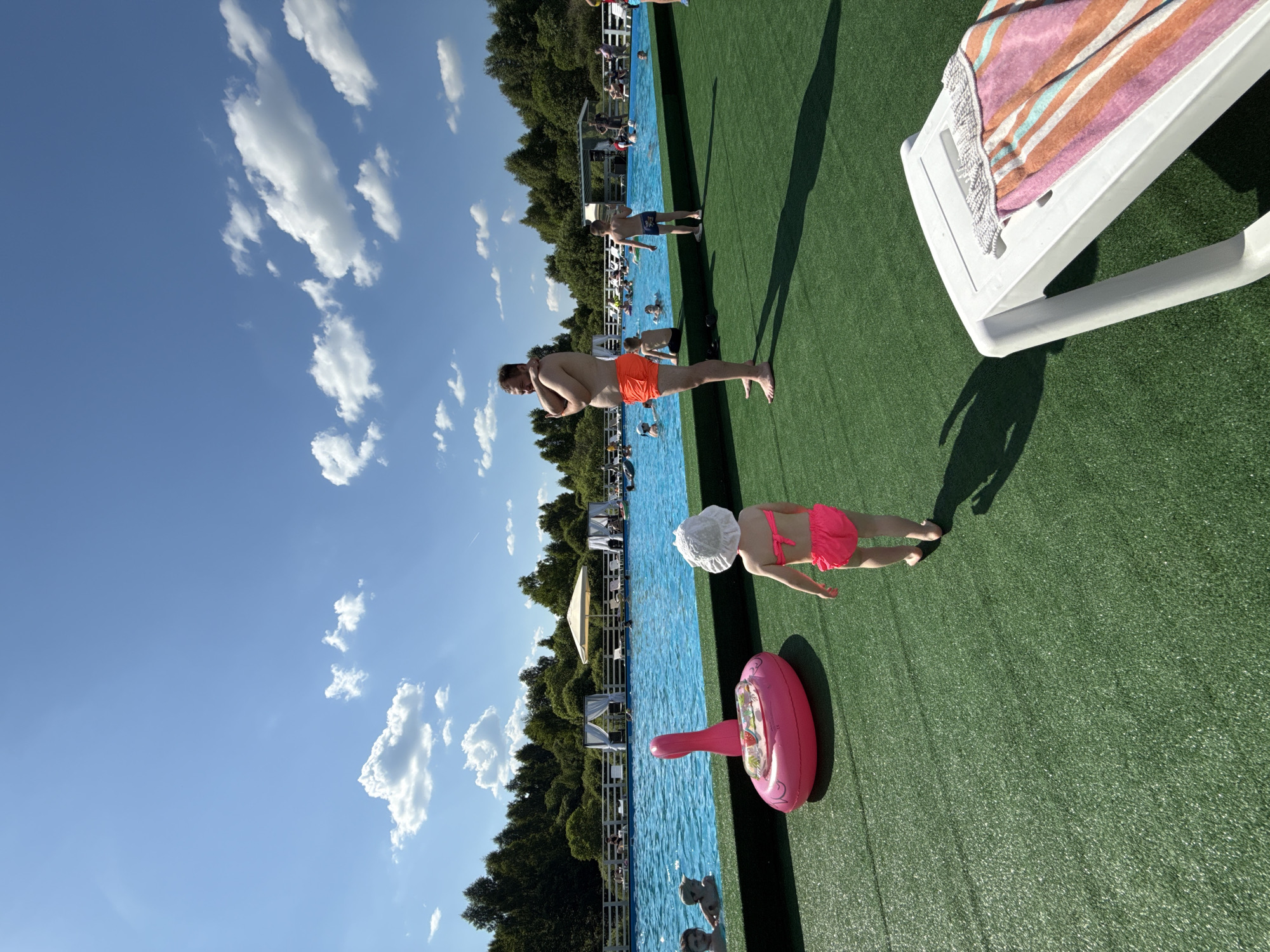 This is a daytime, eye-level, outdoor, full shot photograph taken at a pool facility. The scene depicts a bright, sunny day with a clear blue sky dotted with fluffy white clouds. The foreground is dominated by artificial green turf, upon which a white plastic lounge chair with a striped towel draped over it is visible on the right side. To the left of the chair, a young child wearing a white sun hat and a bright pink bikini is walking away from the camera. In front of the child, a large, inflatable pink flamingo pool float rests on the turf. Beyond the turf is a large, blue swimming pool filled with people. In the background, beyond the pool, is a line of trees and a white fence, suggesting a natural, possibly rural, setting. A few white gazebos with draped curtains and a large cream-colored umbrella are set up near the pool. People are seen swimming, lounging, and socializing in and around the pool. The overall atmosphere is one of leisure and recreation. The location is identified as Koloksha, Russia.